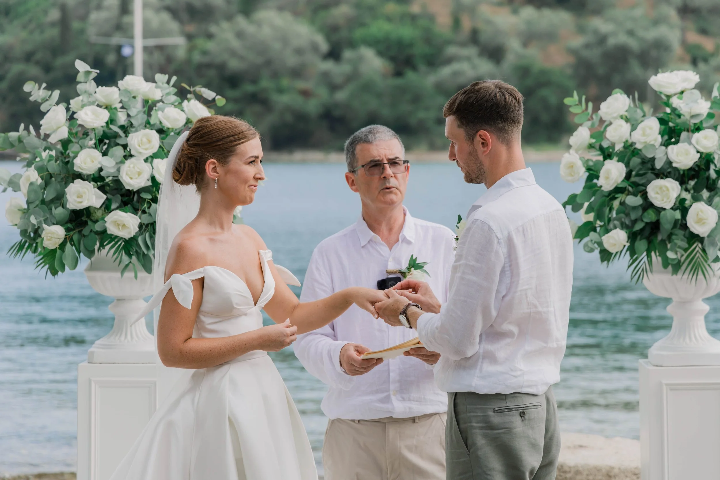 A wedding ceremony by the water with a bride and groom exchanging rings, officiant, and floral arrangements.