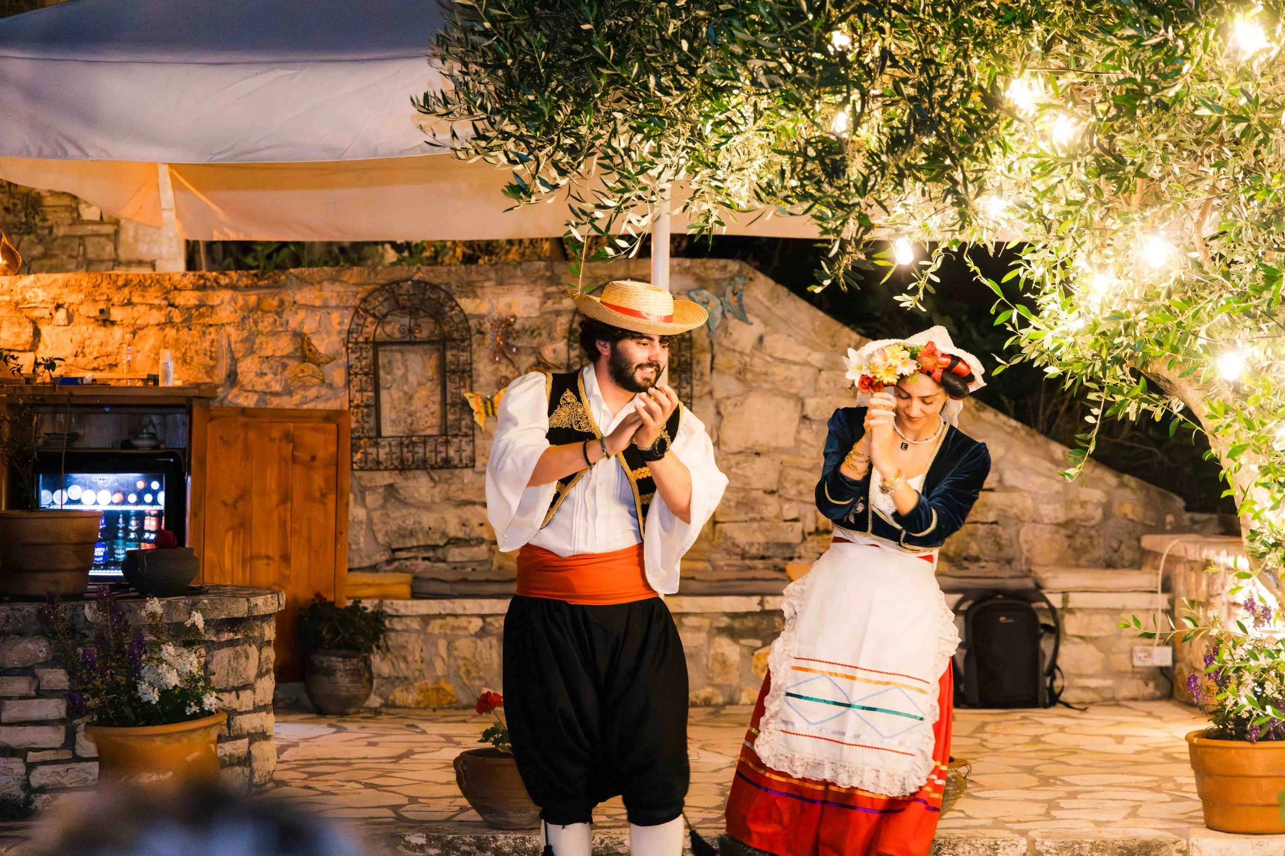Two people in traditional Spanish folk costumes dancing outdoors at night, under a lit tree, with a stone wall in the background.