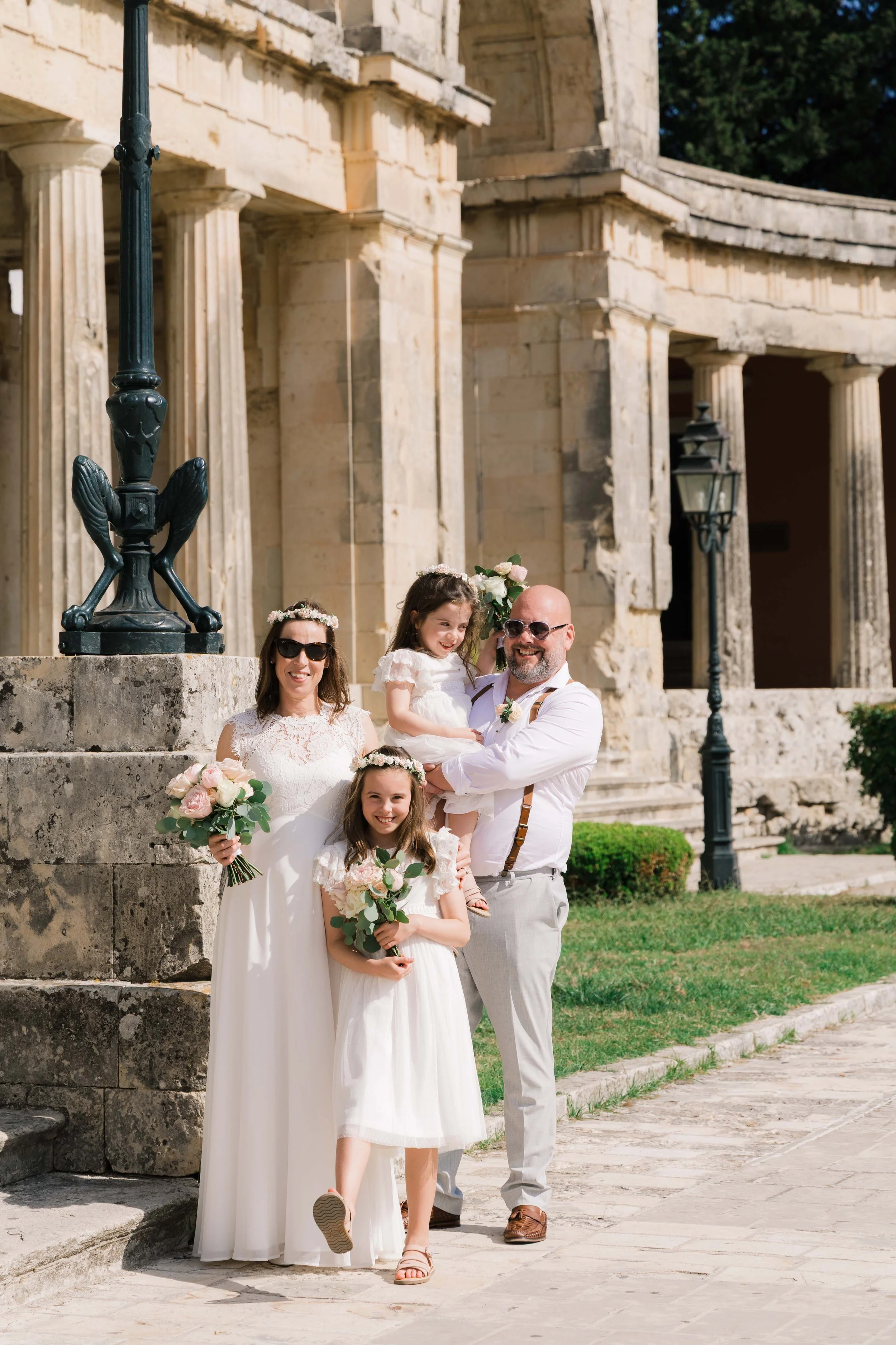 Family celebrating wedding outdoors, standing in front of historic stone building with columns, holding bouquets, smiling.
