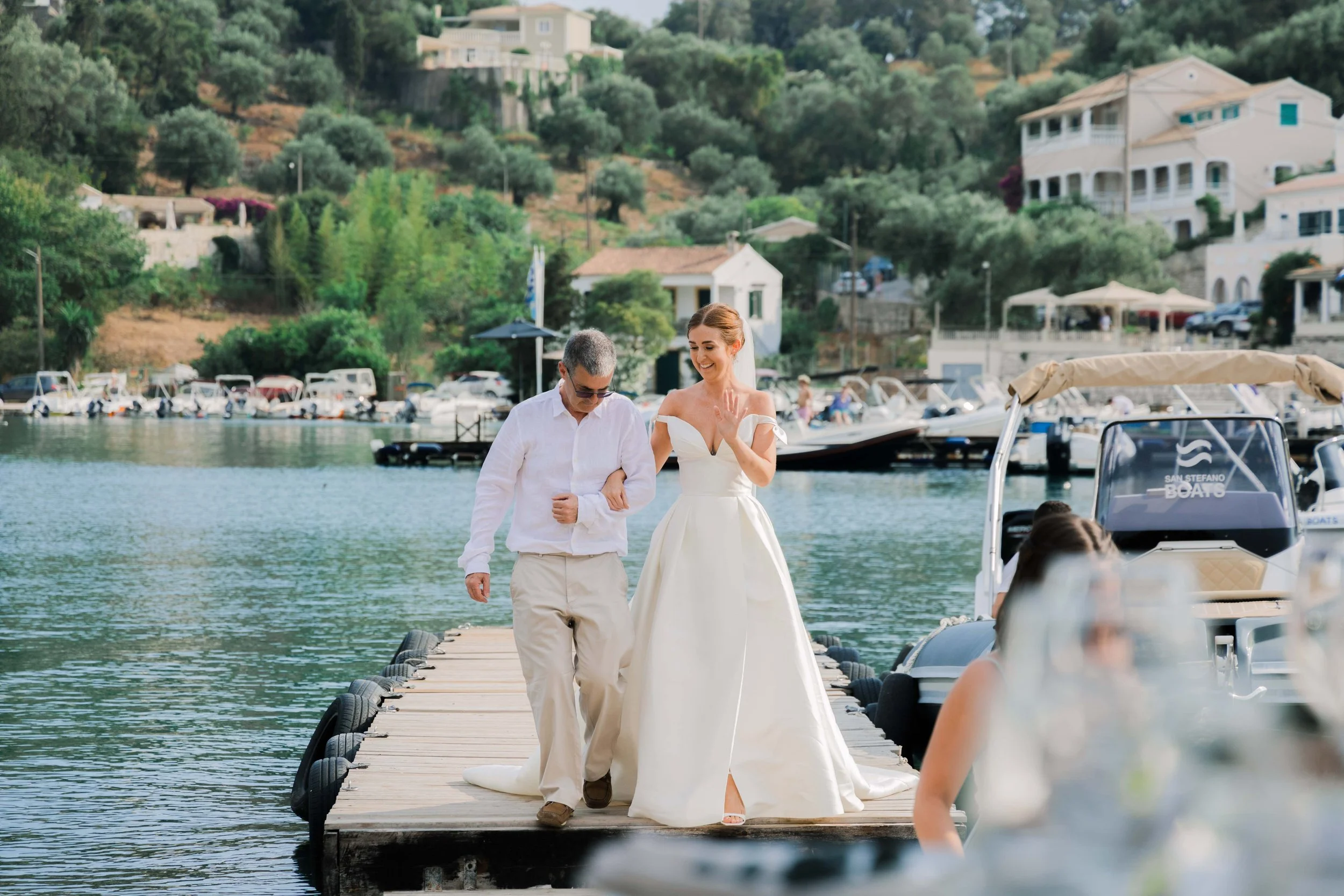 A bride in a white wedding gown walks arm-in-arm with a man in light-colored pants and a white shirt on a dock by the water. There are boats anchored nearby and a hillside with houses and trees in the background.
