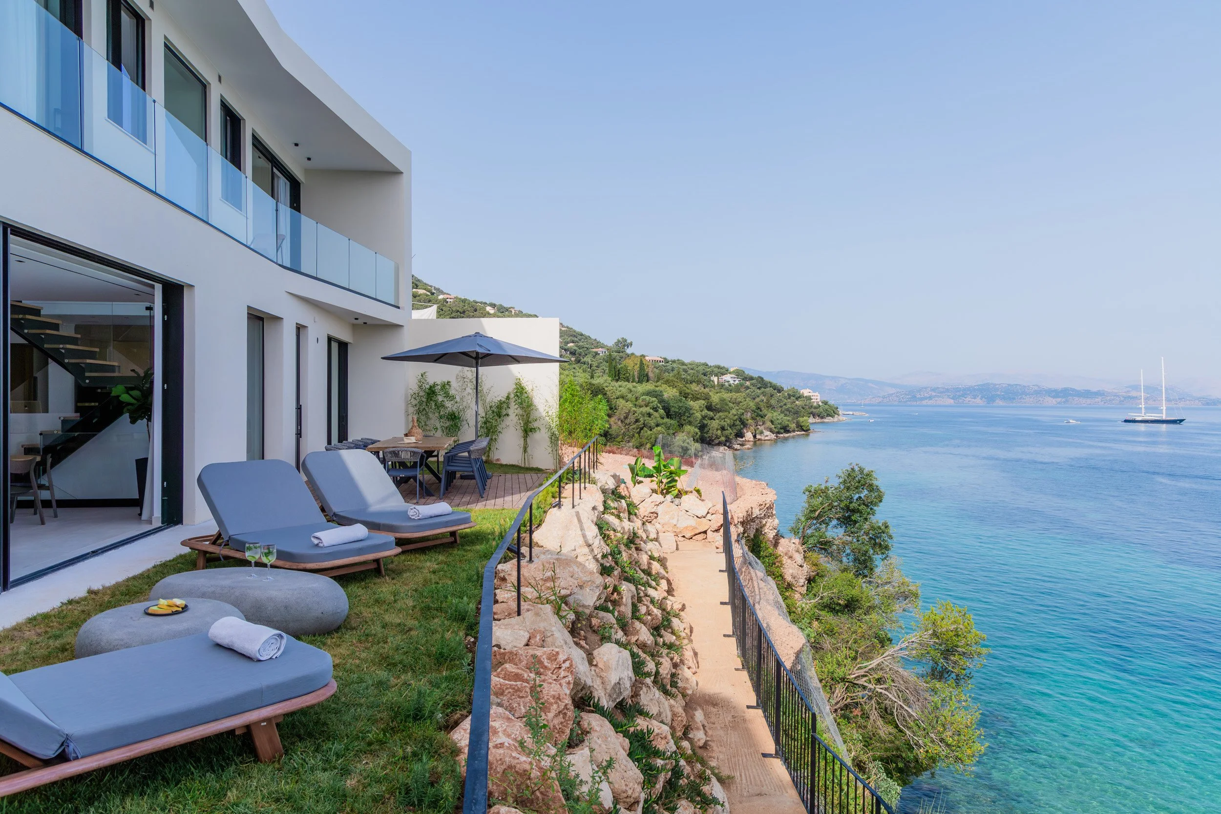 Modern white building with glass balcony overlooking a rocky coastline and calm blue water, featuring lounge chairs, a table with chairs under an umbrella, and a narrow pathway along the rocky edge.