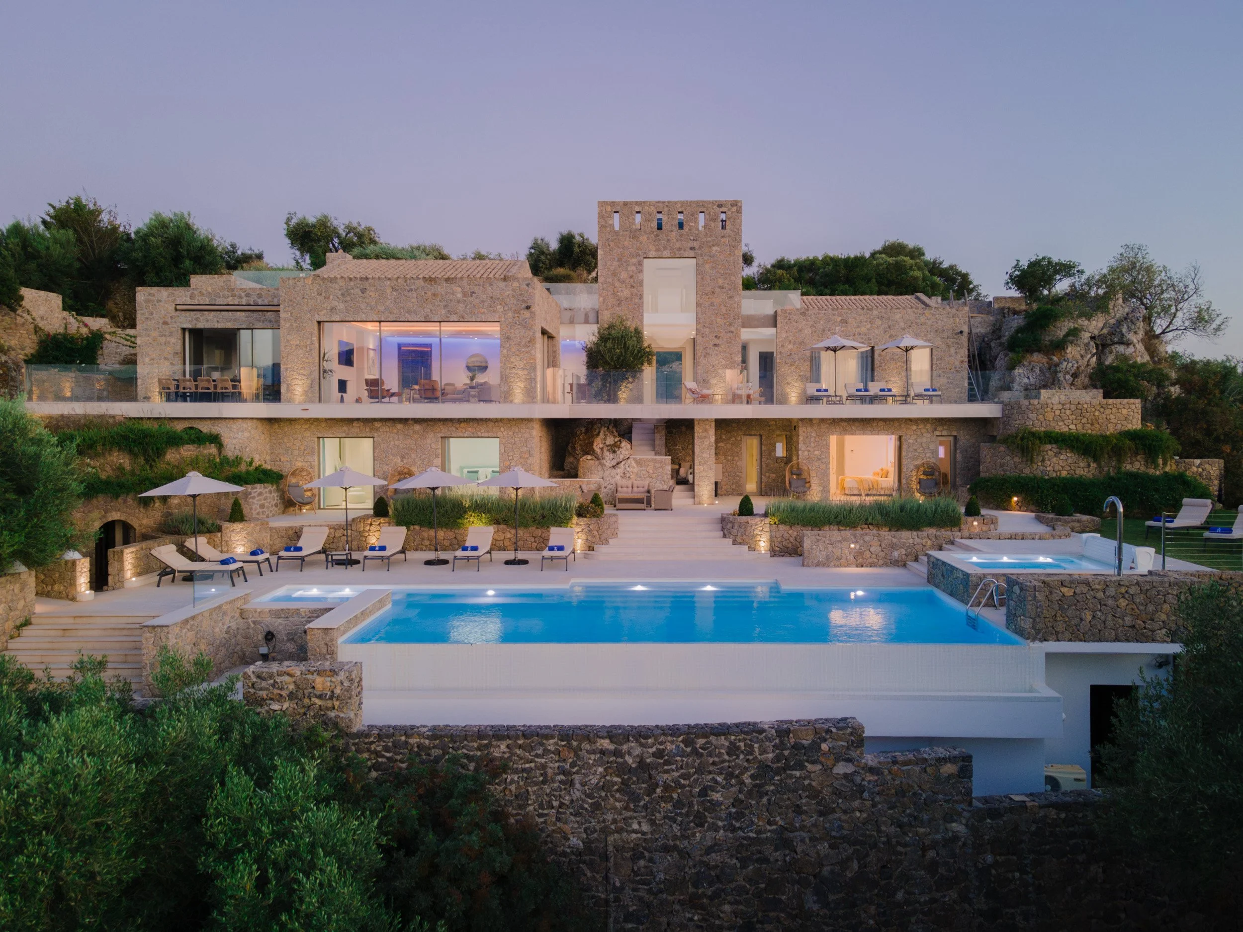 Luxury modern stone house with multiple levels, large glass windows, outdoor pool with lounge chairs and umbrellas, surrounded by greenery at dusk.