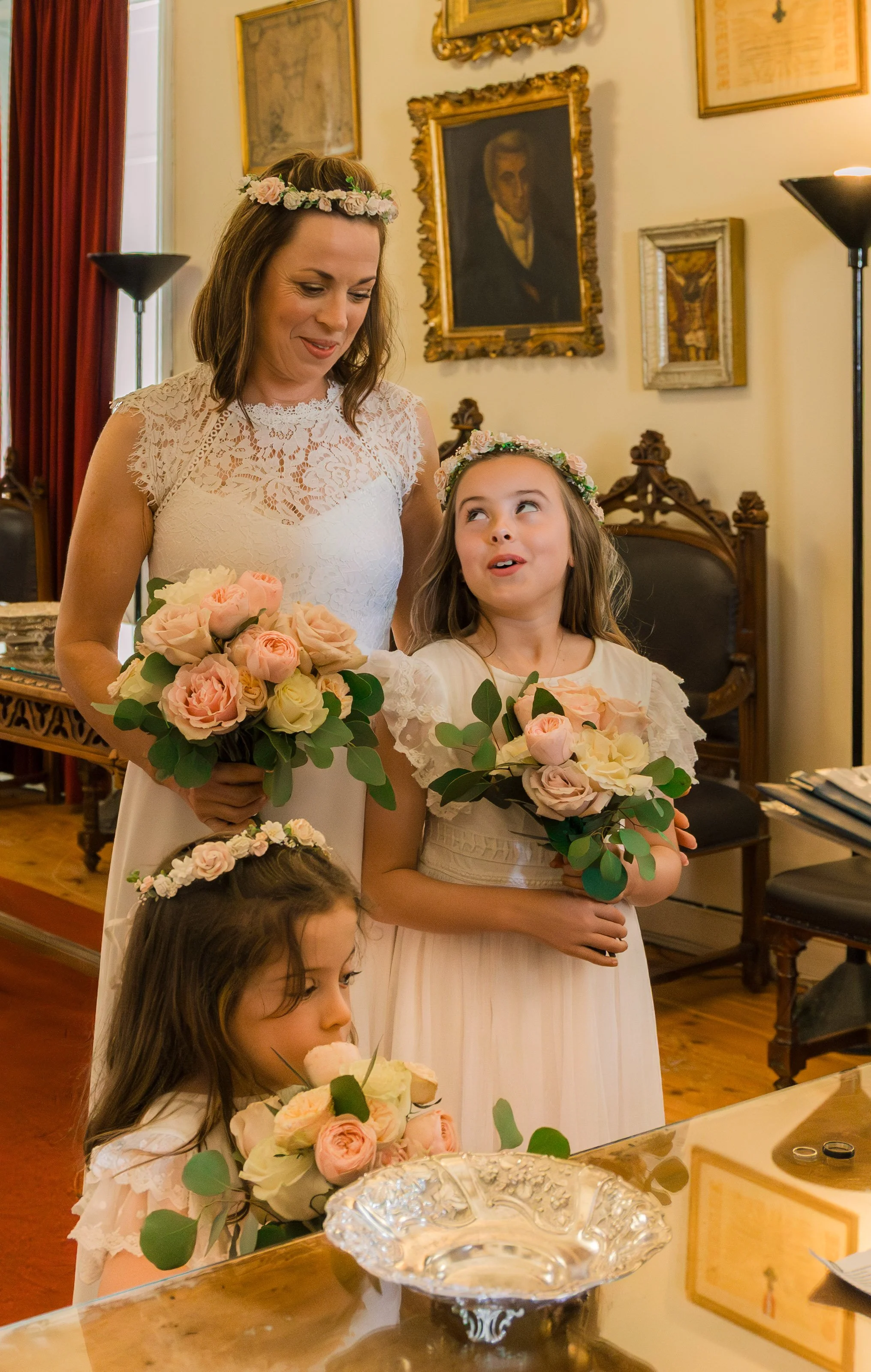 A woman and three young girls dressed in white are standing in a room decorated with framed paintings. The women and girls are holding bouquets of pink and cream roses with green leaves, and all four have flower crowns on their heads. The women and g
