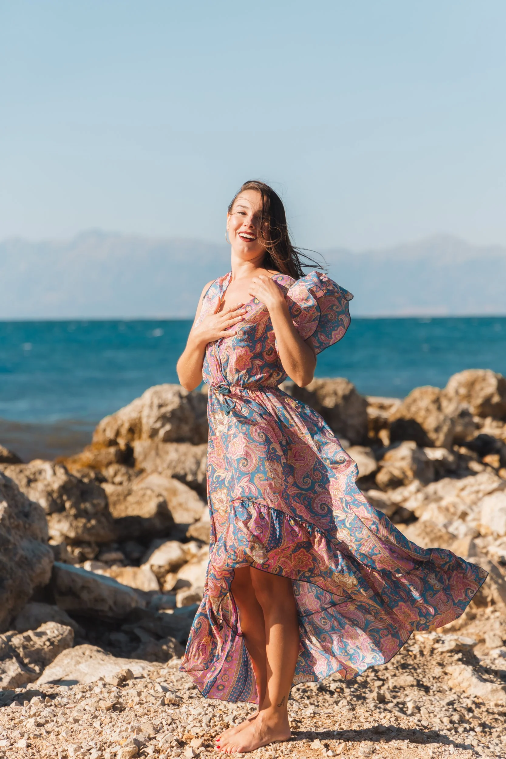 A woman standing on rocky beach near the ocean, wearing a colorful, flowing patterned dress, smiling, with her hand on her chest.