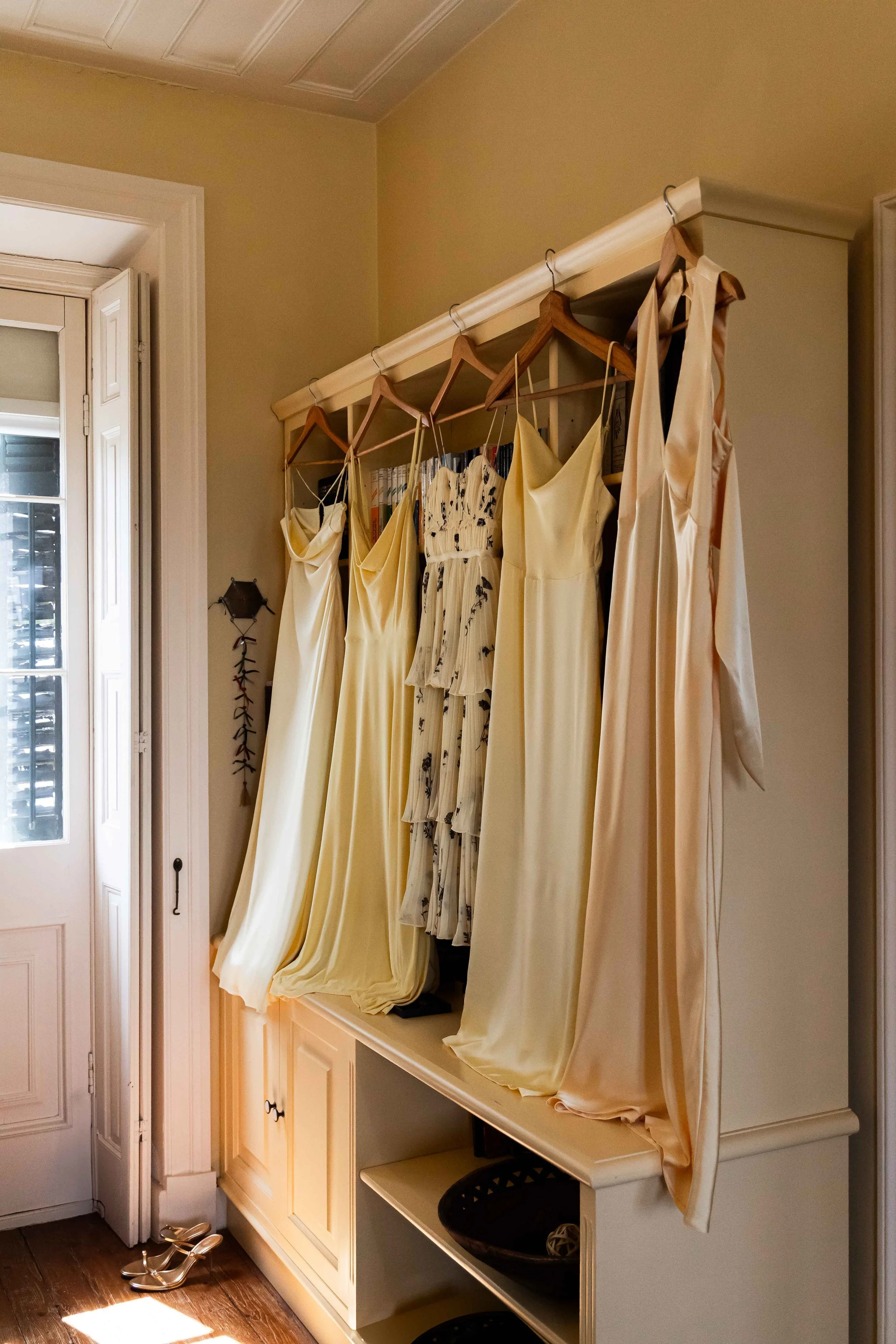 A closet with six hanging dresses, mostly cream and patterned, on a white rack near a window with shutters. Shoes are on the floor near the door.