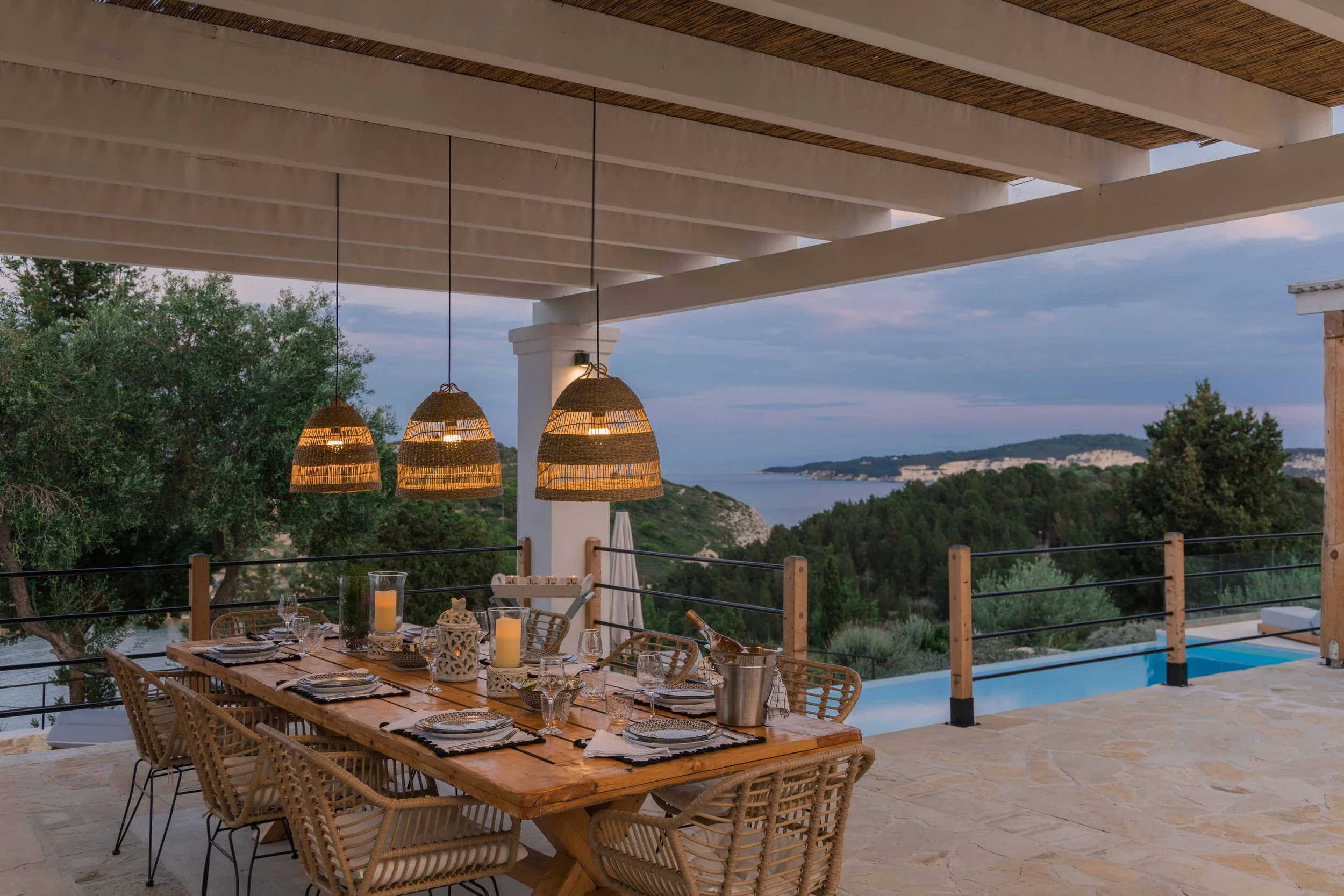 An outdoor dining area on a patio with a wooden table set for a meal, surrounded by wicker chairs. Hanging from the ceiling are three woven pendant lights. The backdrop includes green trees, rolling hills, and a distant view of the sea or a large lak