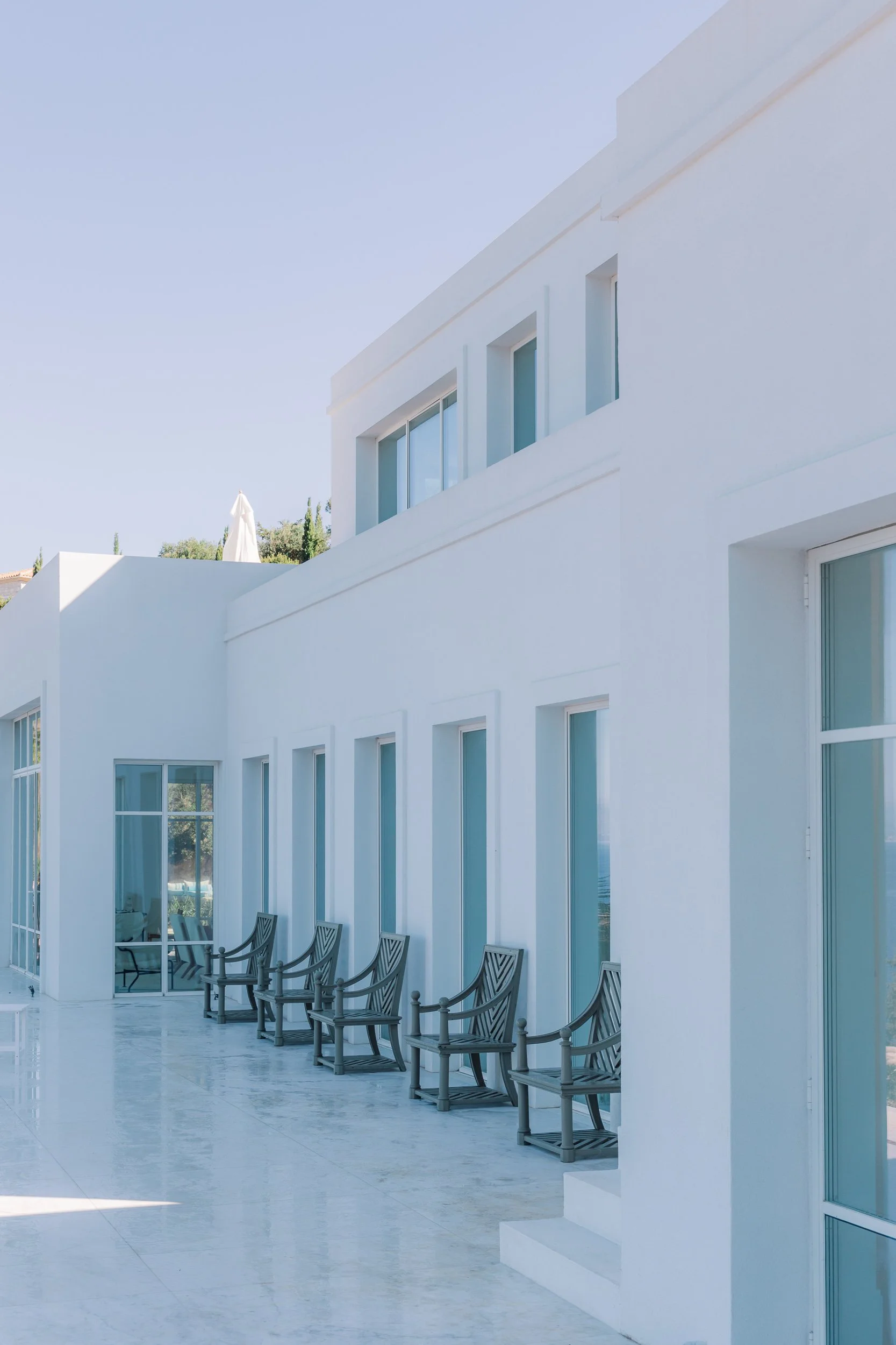 White modern building with large glass windows and black chairs on a patio under a clear blue sky.