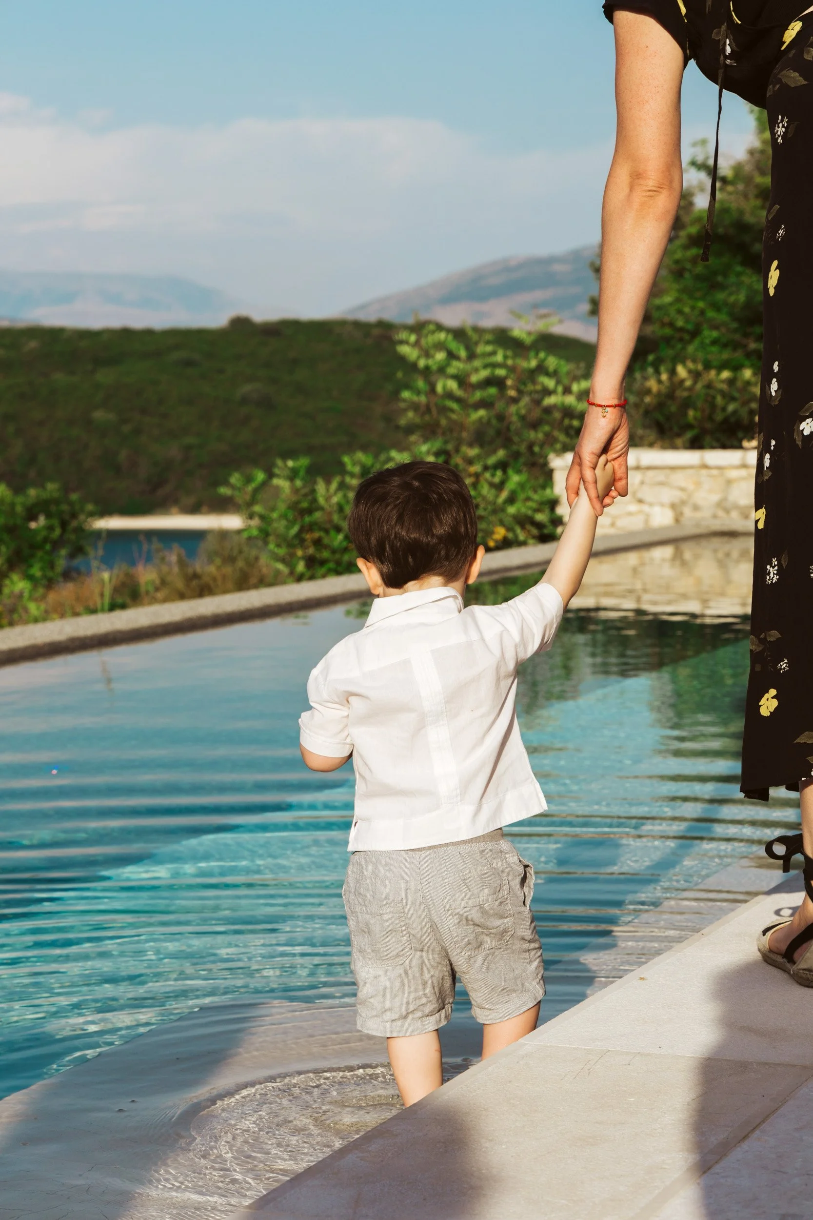 A young boy is standing at the edge of a pool, being held by an adult woman, possibly his mother, by the hand. The background shows a scenic view of green hills and mountains under a partly cloudy sky.