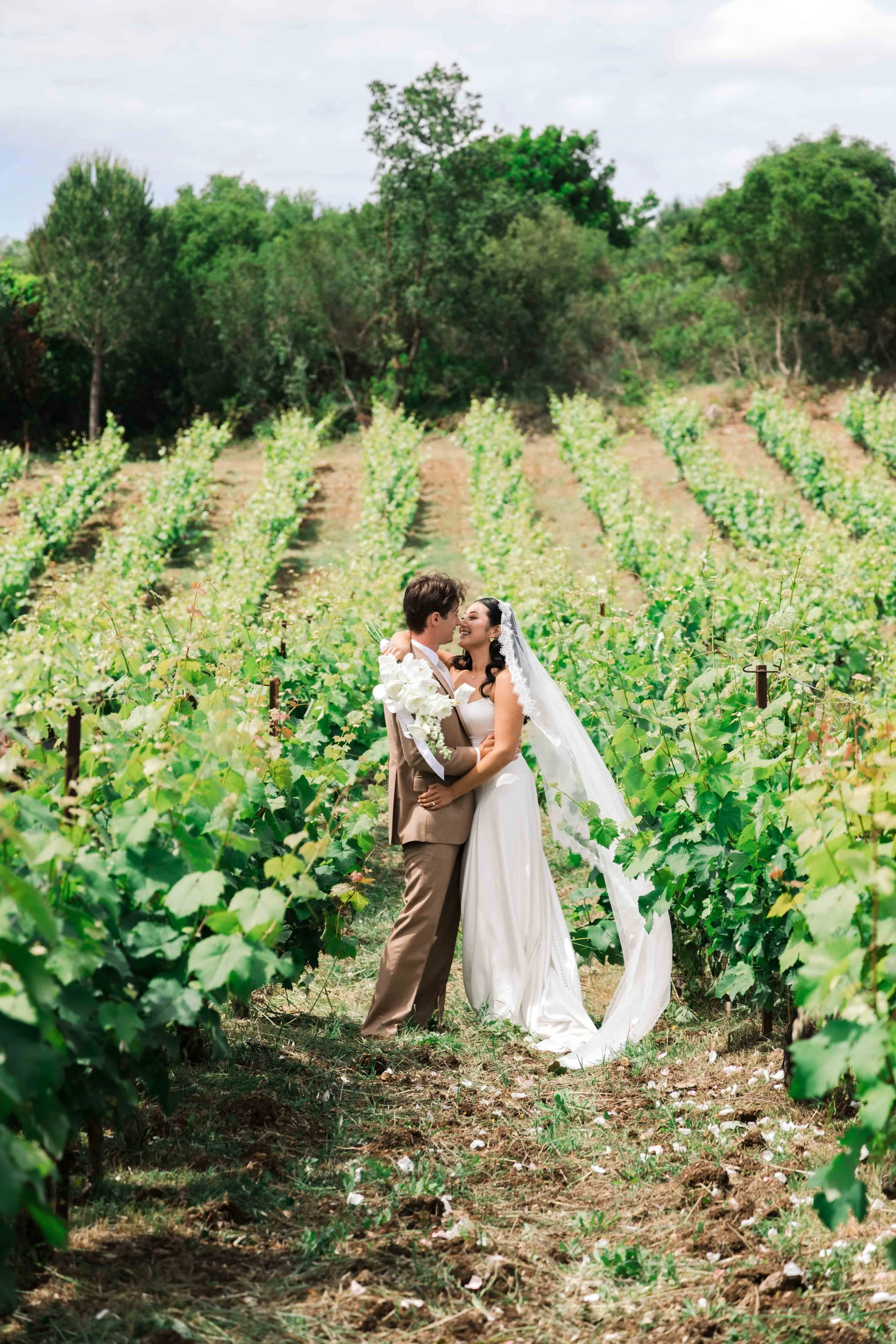 A newlywed couple in wedding attire in a vineyard, embracing and smiling at each other.