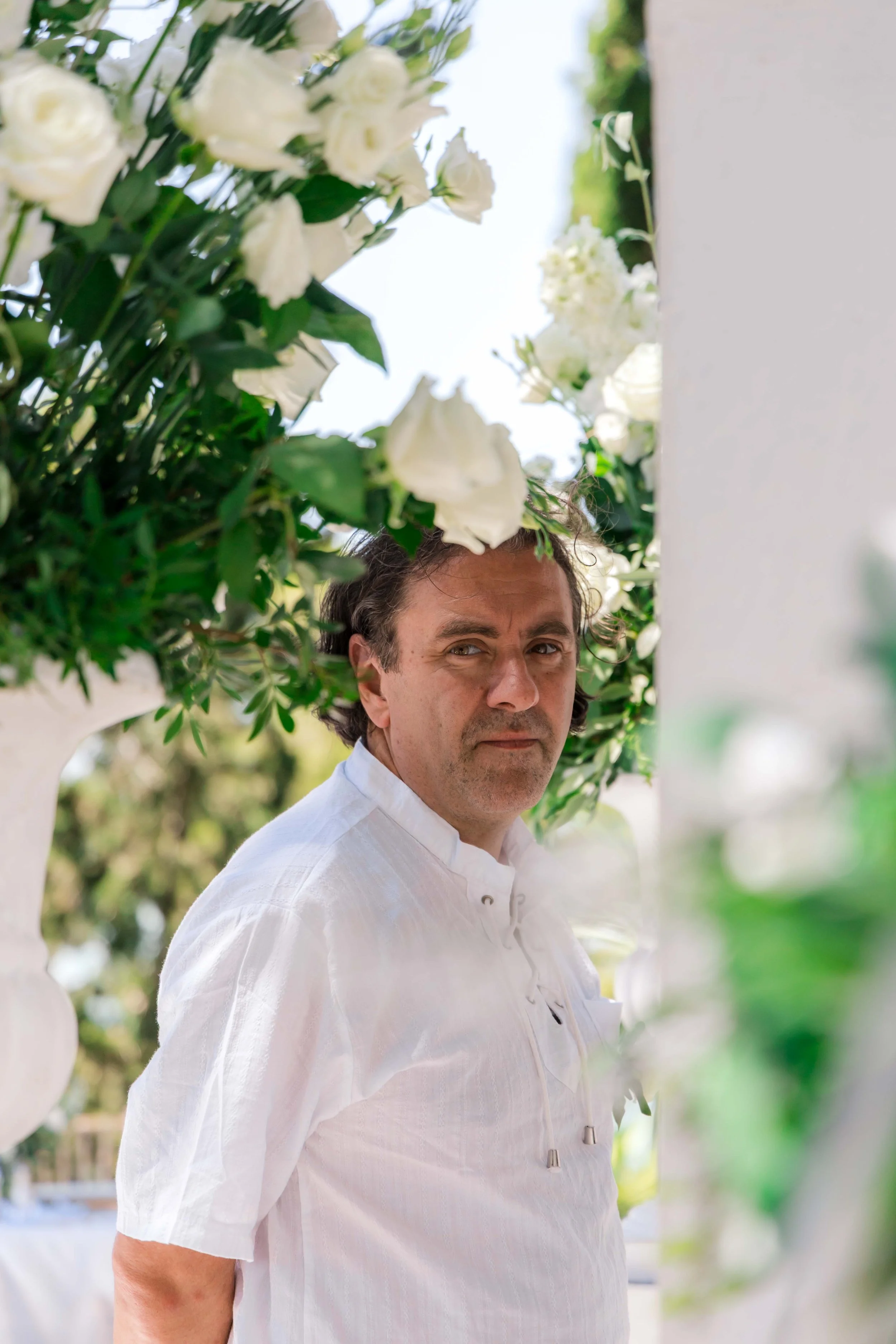 A man in a white shirt standing among white flowers, looking directly at the camera, outdoors with a blurred background.
