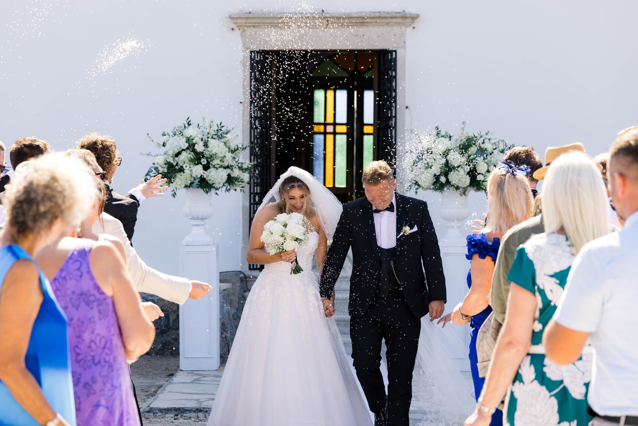 A bride and groom holding hands and smiling as they walk out of a church, surrounded by guests throwing confetti, with large floral arrangements beside the church entrance.