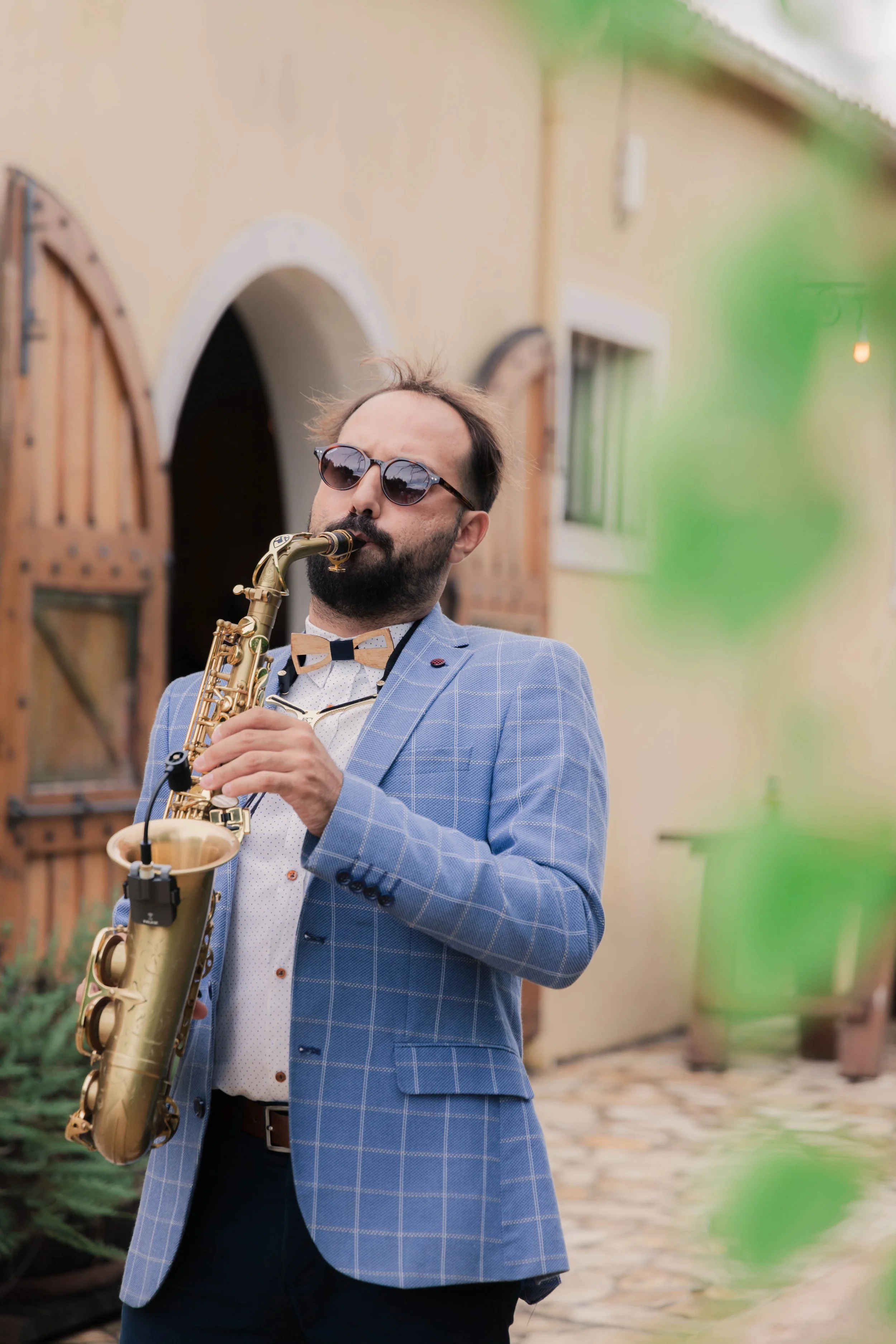 A man wearing sunglasses and a blue checkered blazer is playing a saxophone outdoors near a building with wooden windows and a stone pathway.