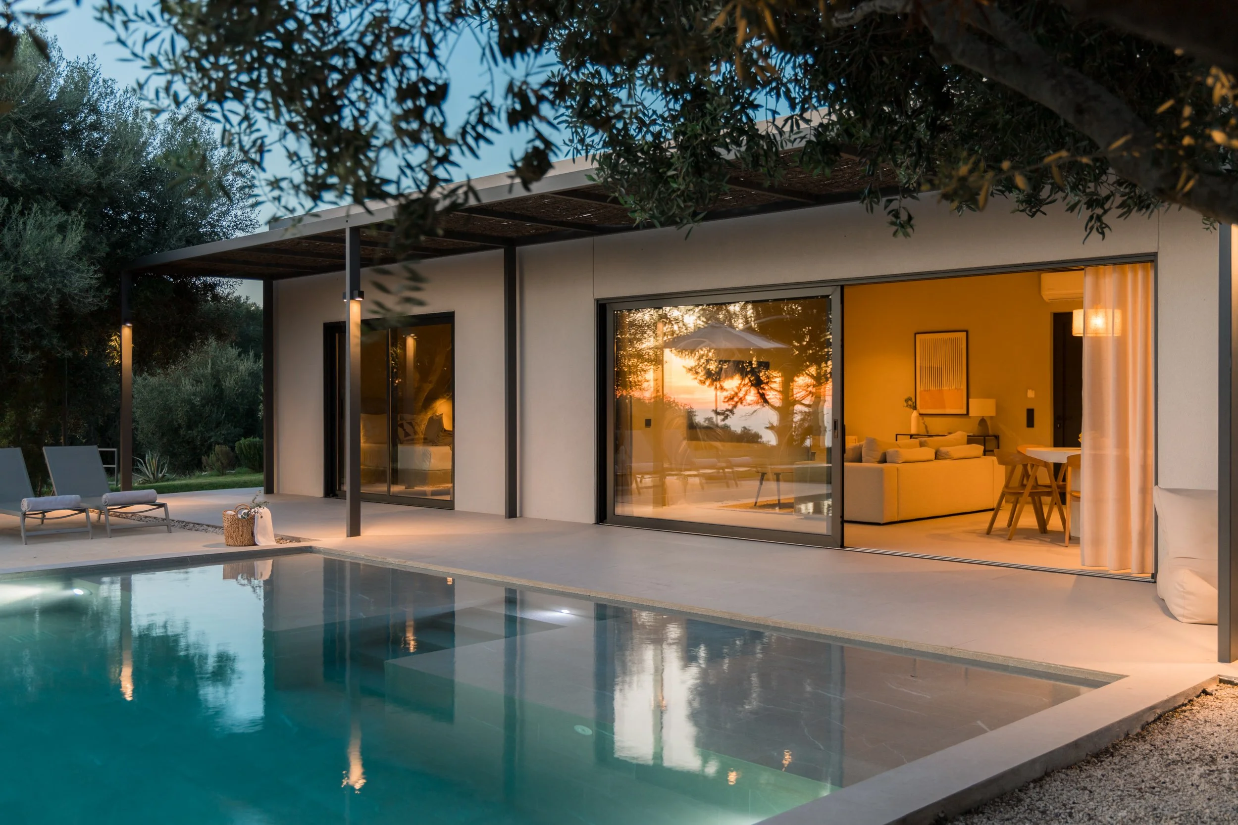 Modern house with large sliding glass doors, illuminated interior, outdoor pool area, seating, and surrounding trees at sunset.