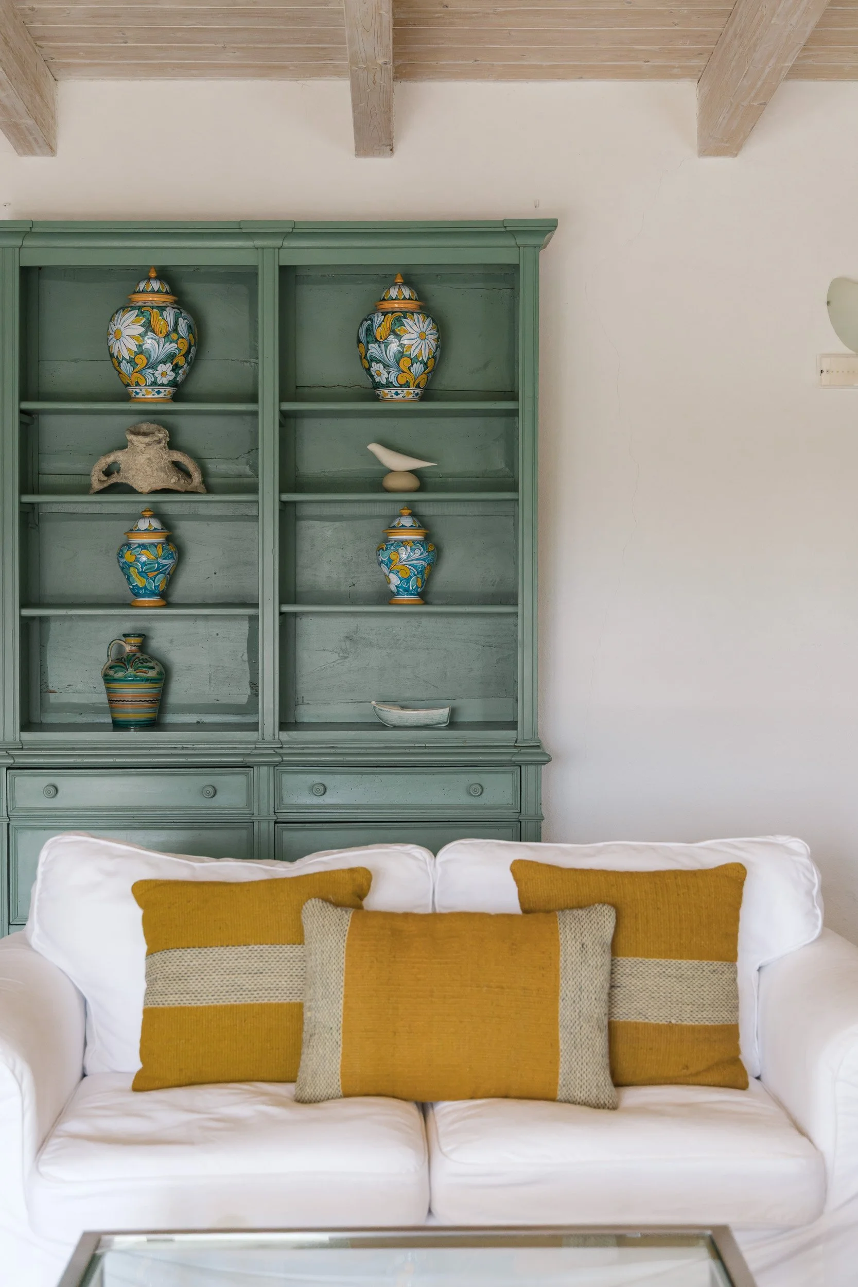 A living room with a green wooden display cabinet filled with decorative vases and sculptures, and a white sofa with three mustard-colored pillows in front of it.