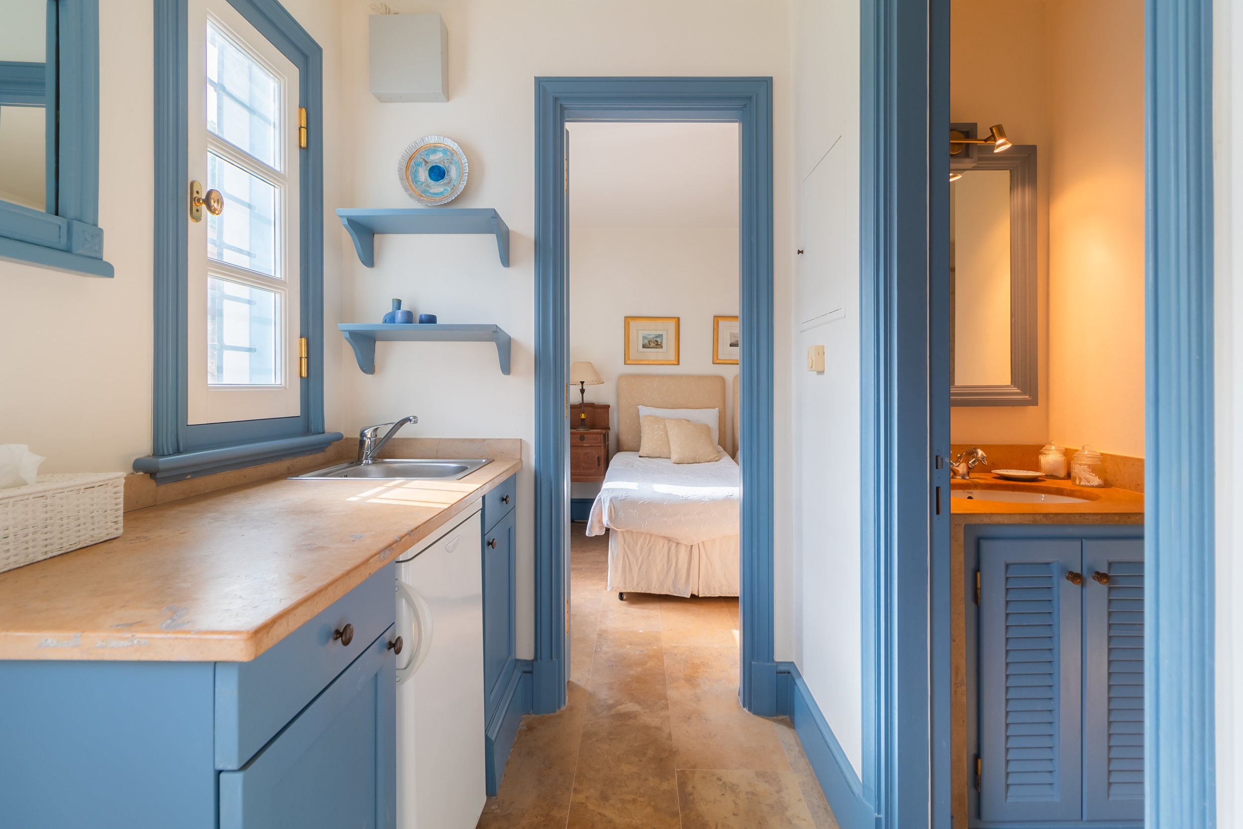 View of a cottage interior showing a small kitchen with blue cabinets and trim, a countertop with a sink, a window, a bedroom with a white bed and pillows, and a bathroom with a mirror and sink.