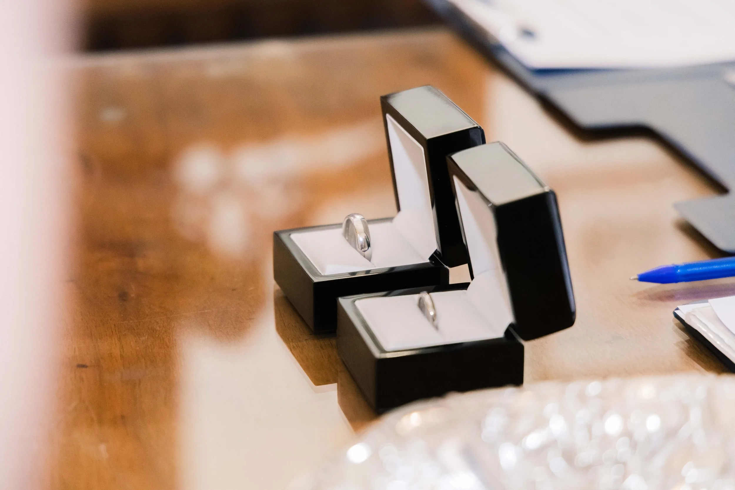 Two black ring boxes on a wooden table, each containing a wedding band, with a blue pen and a laptop in the background.