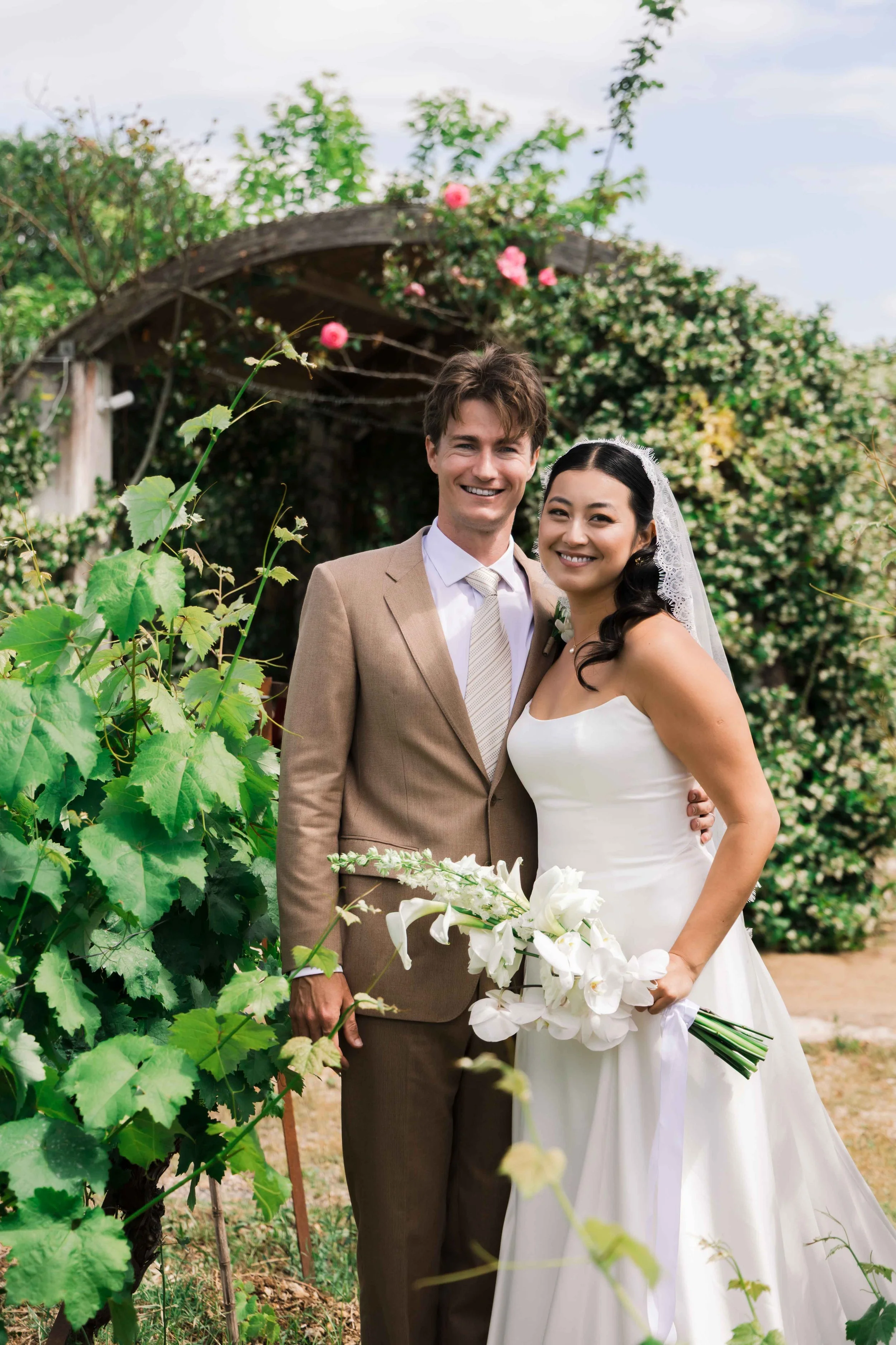A happy couple in wedding attire standing outdoors, surrounded by greenery and flowering plants, on their wedding day.
