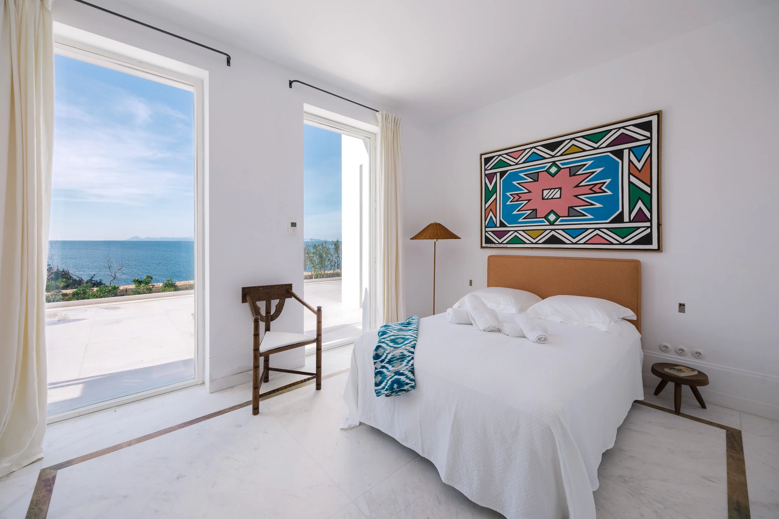 A bright hotel room with a large window and a sliding door showing ocean views. The circular bed has white linens and pillows, a tan headboard, and a blue patterned throw. There is a wooden chair, a small side table, and a colorful abstract artwork a