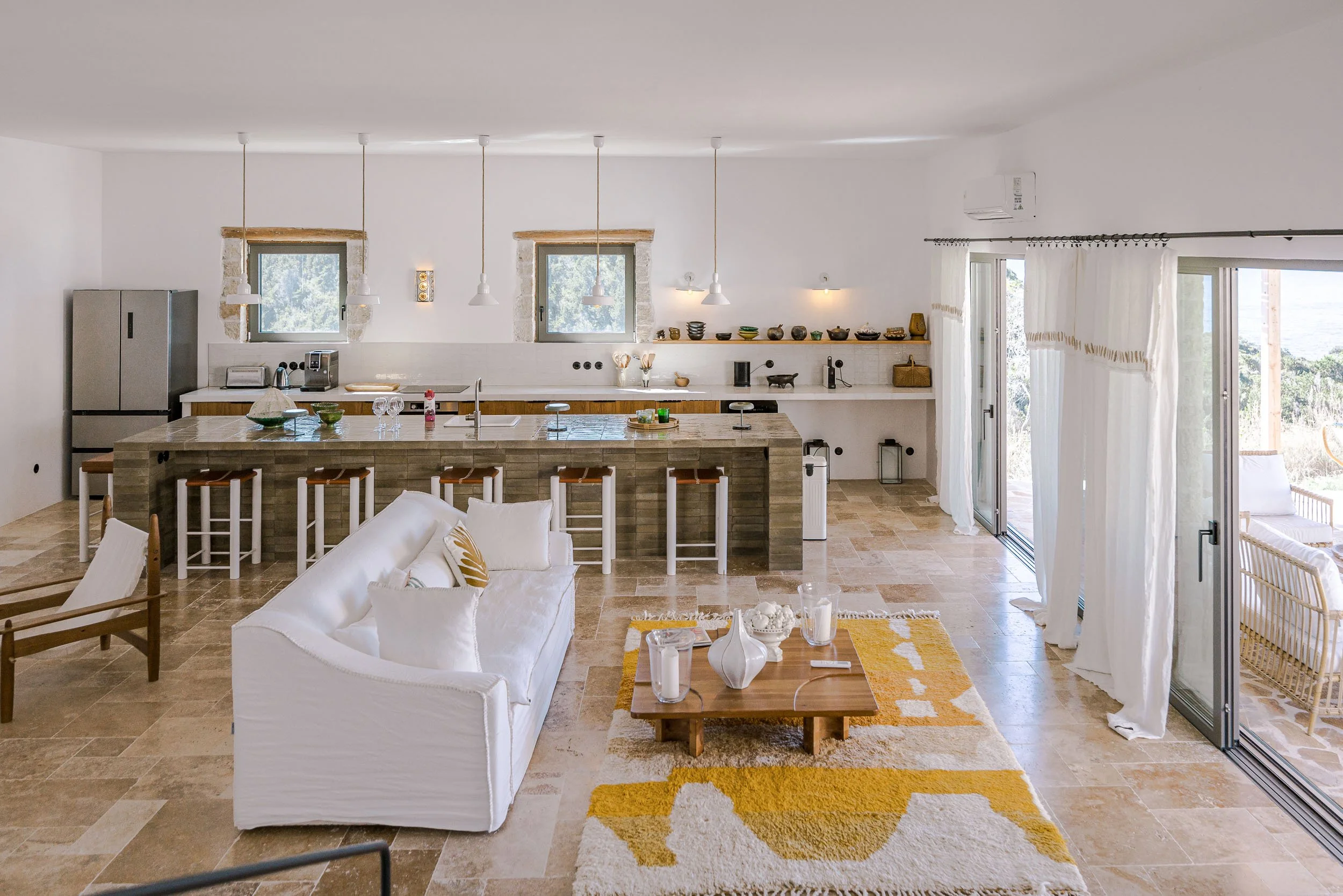 Open-concept kitchen and living area with white walls, beige tiled floor, white curtains, and sliding glass doors leading outside. The kitchen features two small windows, pendant lighting over a large central island with barstools, and open shelves w