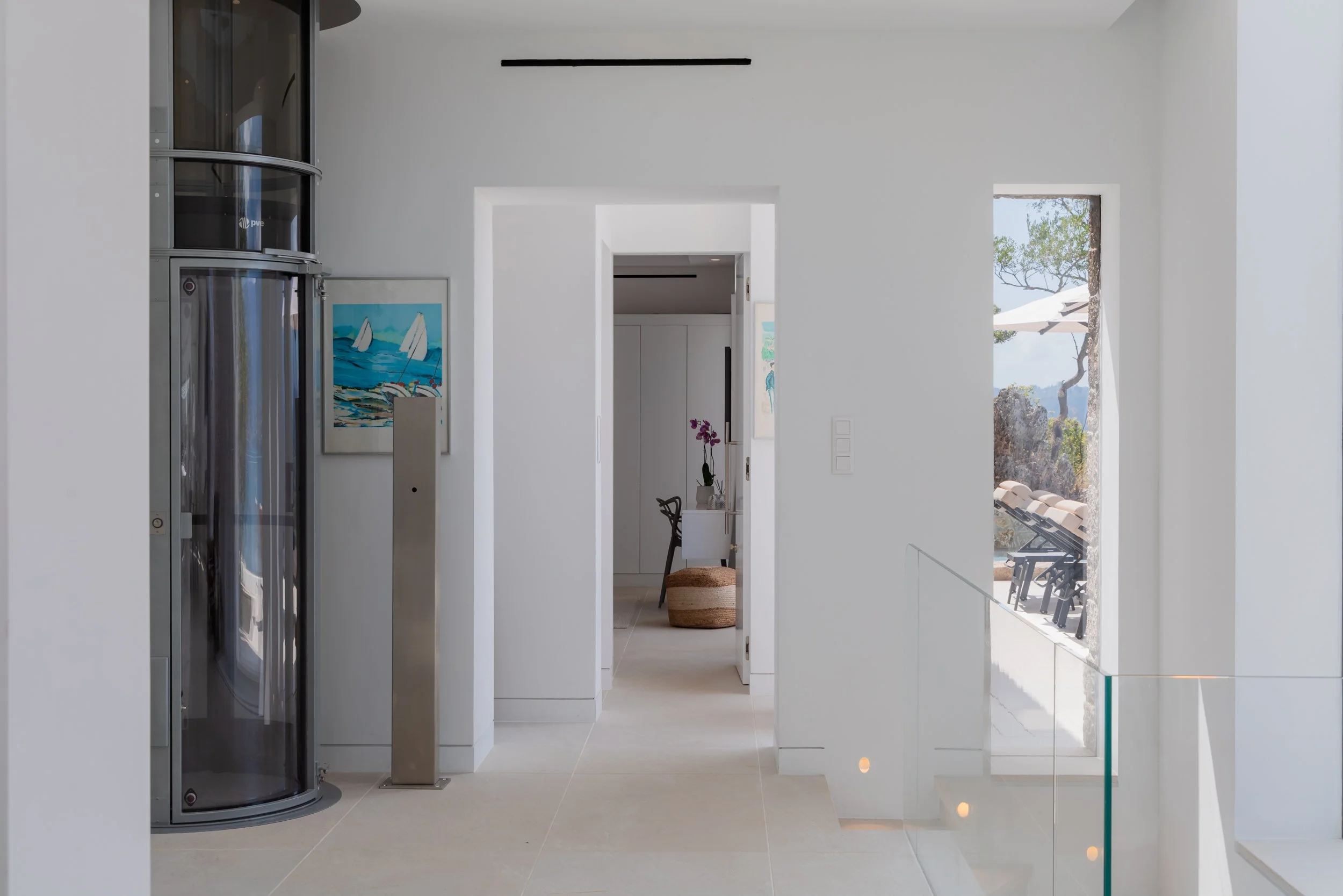 Minimalist interior with white walls, with visible doorway leading to a room with a white cabinet and a purple orchid plant. There's a window showing outdoor lounge chairs and trees.