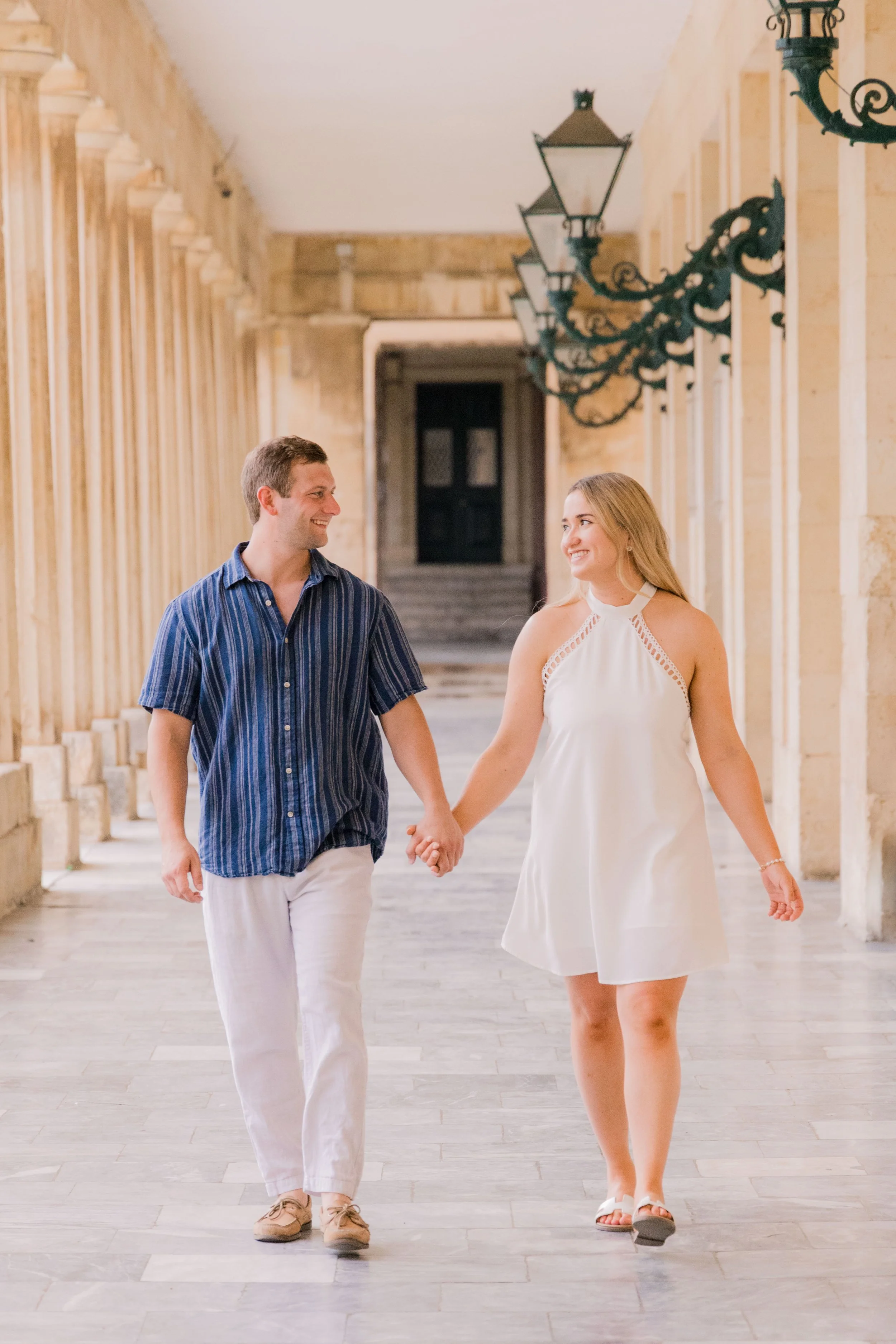 A young couple holding hands and walking through a covered outdoor corridor with lantern-style lights and stone columns, smiling at each other.
