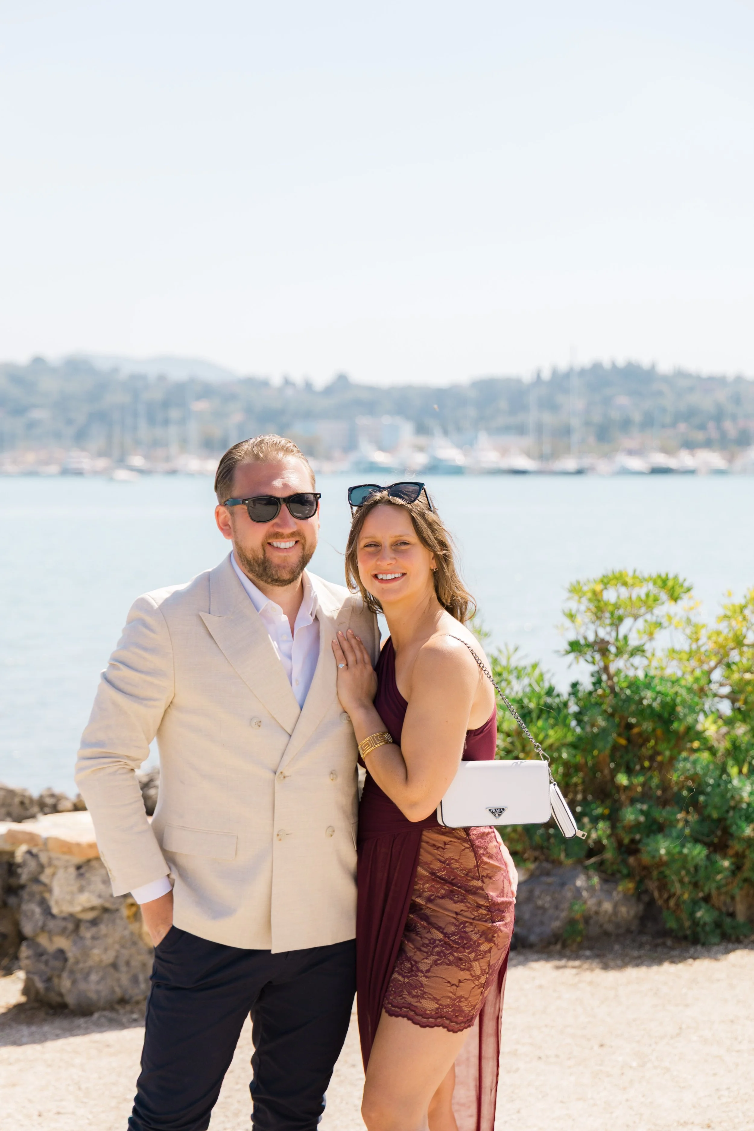 A smiling man and woman standing outdoors near a body of water, with boats and hills in the background. The man is wearing a light-colored suit jacket and sunglasses, and the woman is wearing a burgundy dress with lace details and sunglasses on her h
