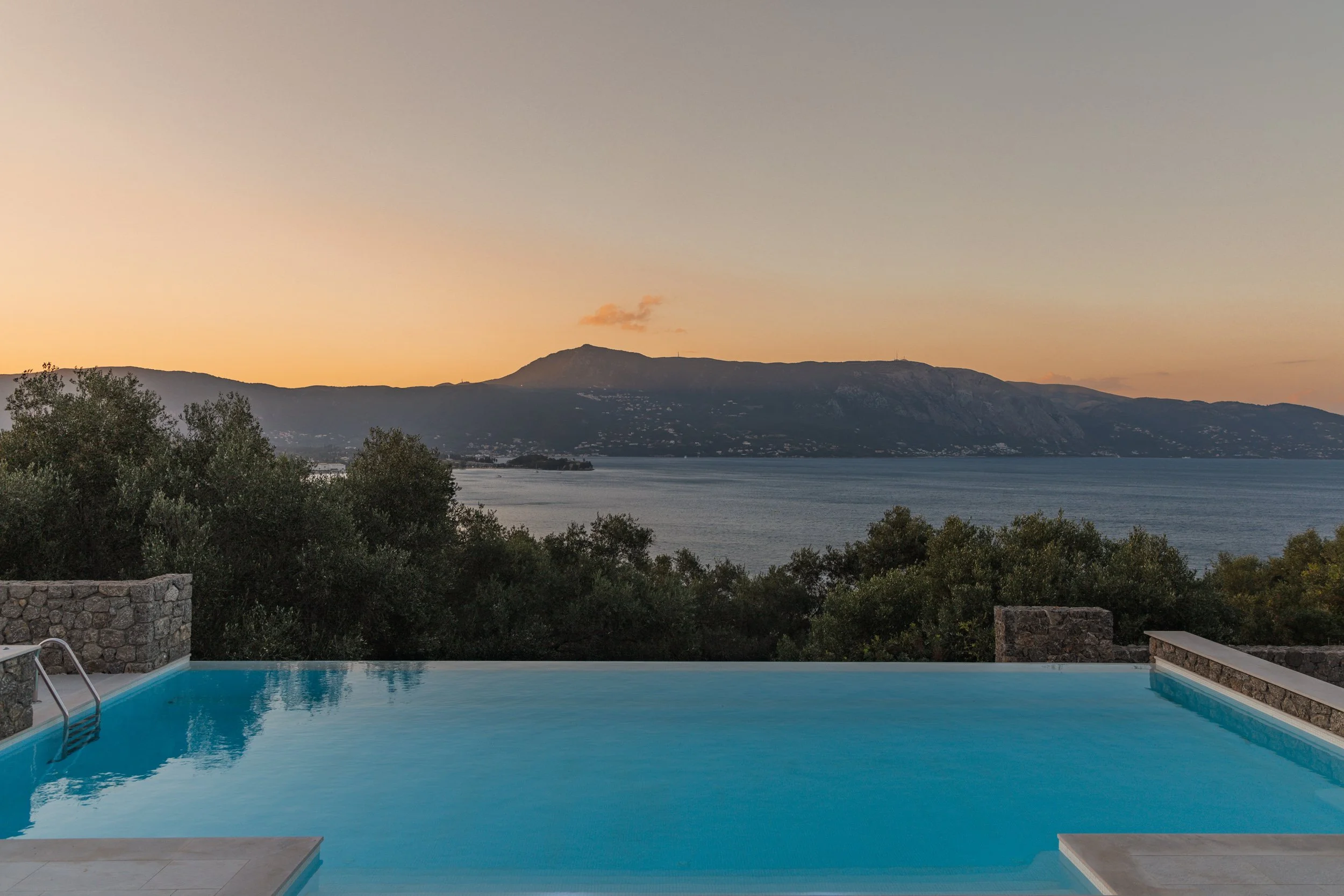 Swimming pool overlooking a lake or sea with mountains in the background during sunset.
