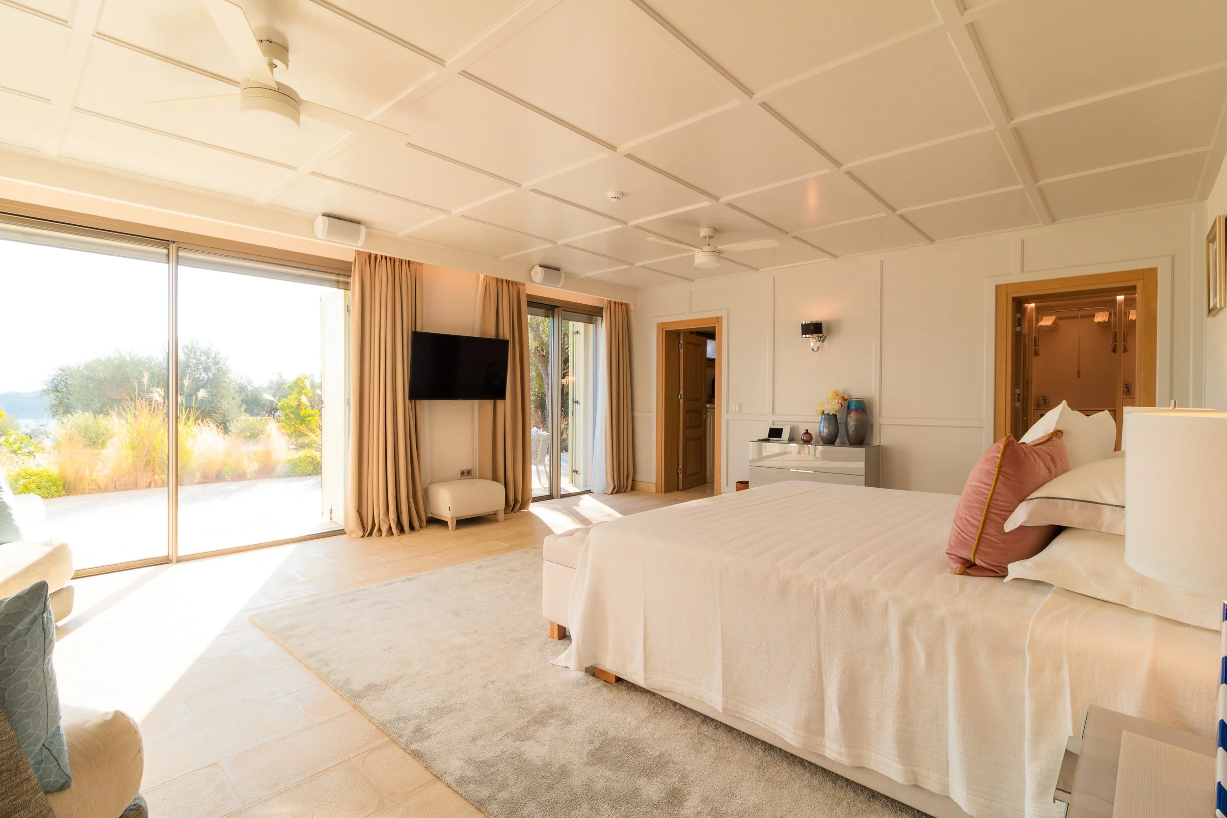 Bright, spacious bedroom with a large bed, sliding glass doors with curtains leading to an outdoor area, a wall-mounted TV, and minimal decor.