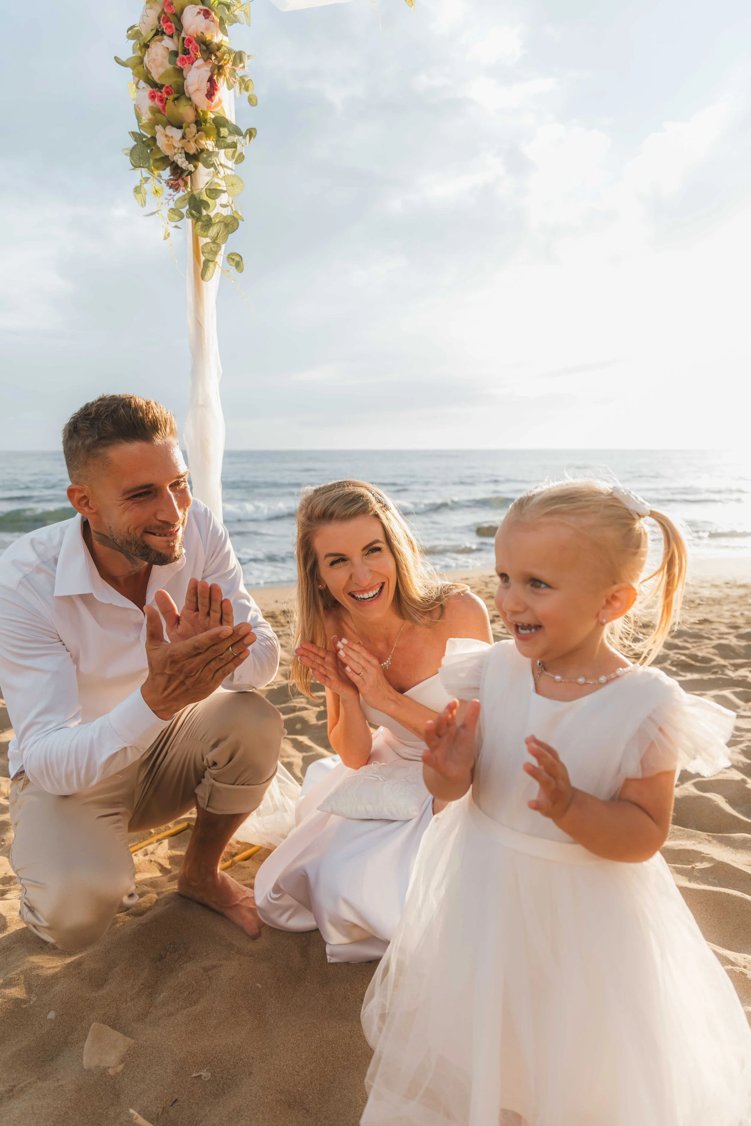 A family celebrating at the beach during sunset, with a decorated archway in the background, everyone dressed in white, smiling, and clapping.