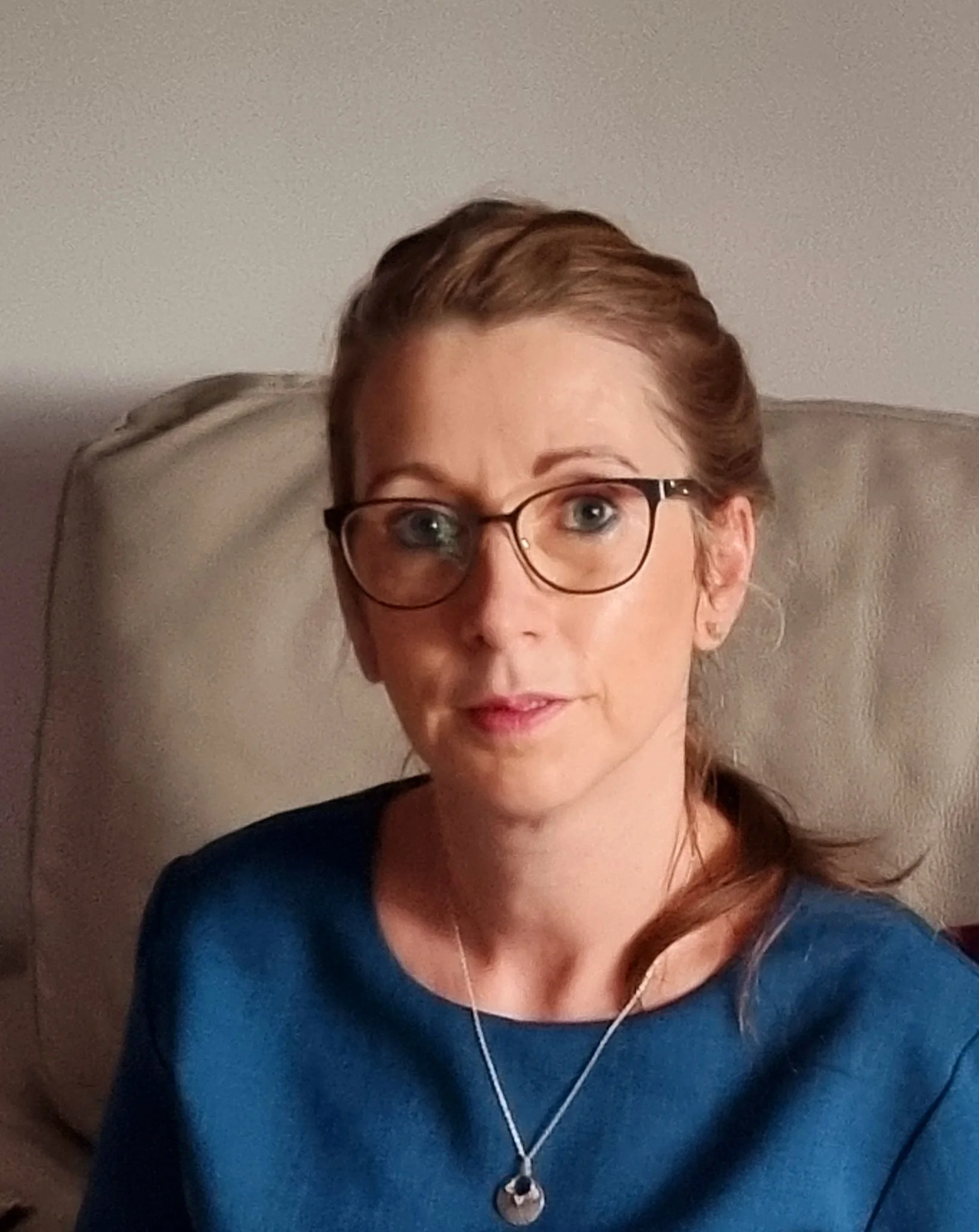 A woman with red hair and glasses sitting on a beige chair in front of a plain light-colored wall.