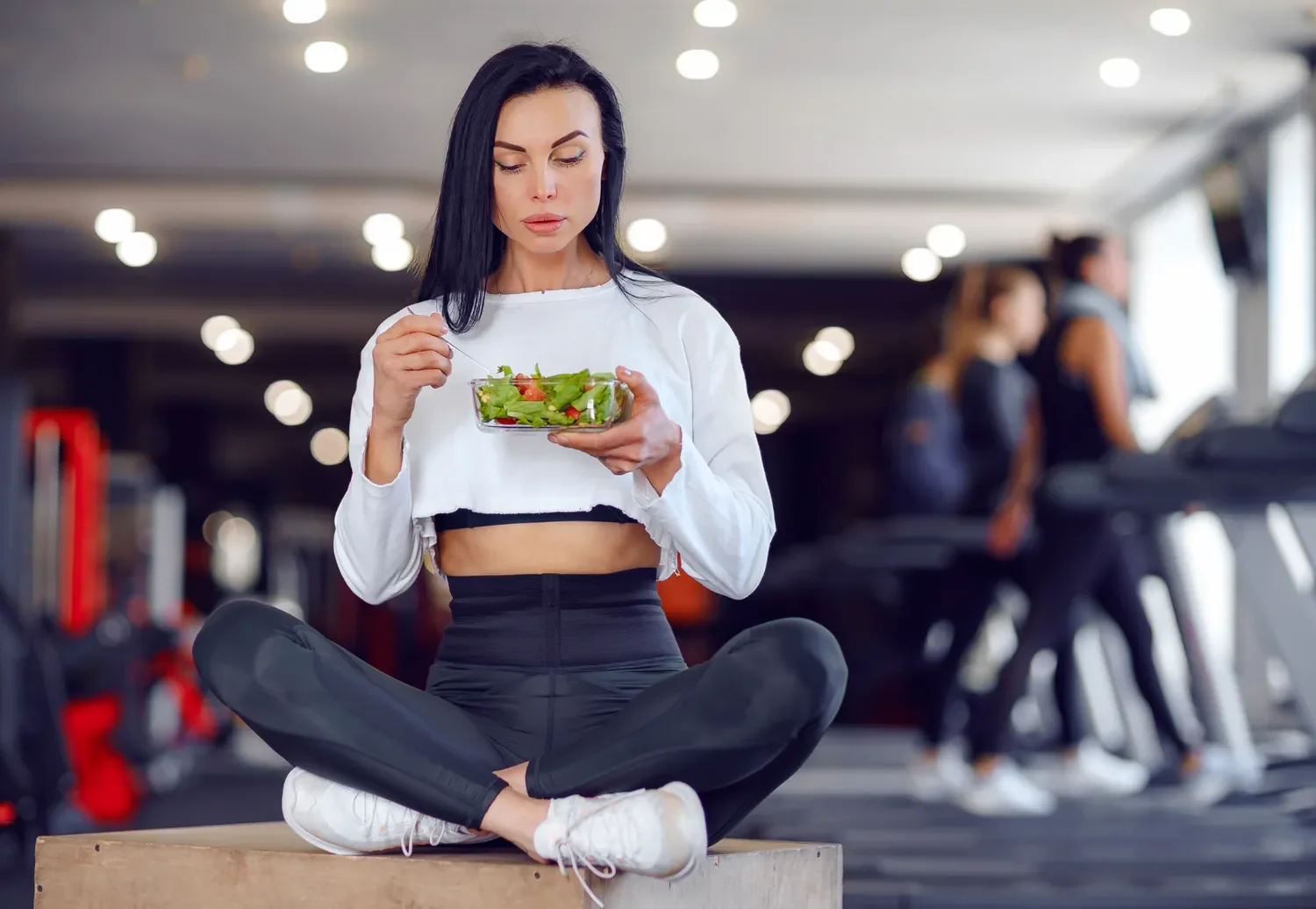 woman sitting cross legged in a gym eating a salad