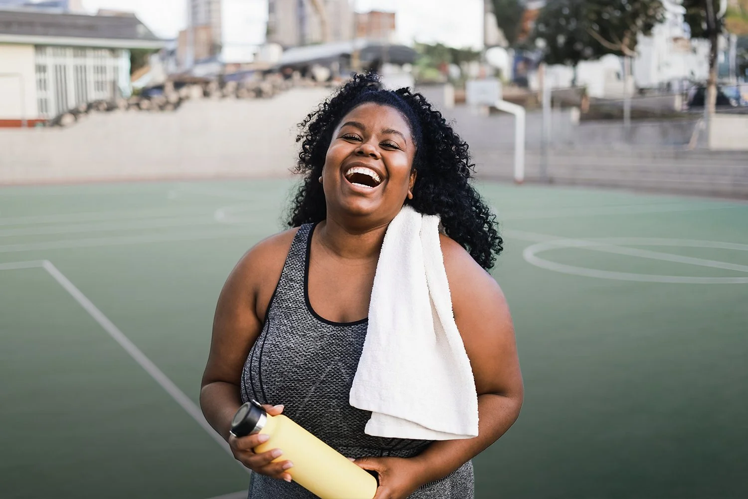 A smiling woman with curly hair stands on an outdoor basketball court after a workout, holding a yellow water bottle with a white towel draped over her shoulder