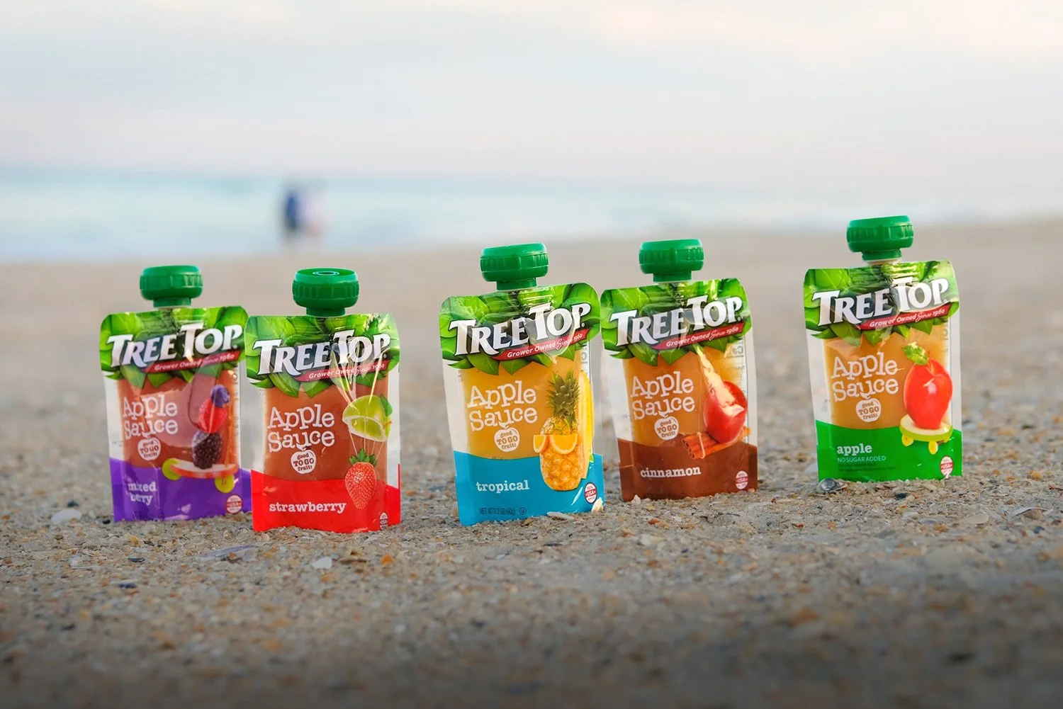 Five different flavors of Tree Top Apple Sauce pouches—Mixed Berry, Strawberry, Tropical, Cinnamon, and Apple—lined up on a sandy beach with the ocean in the background
