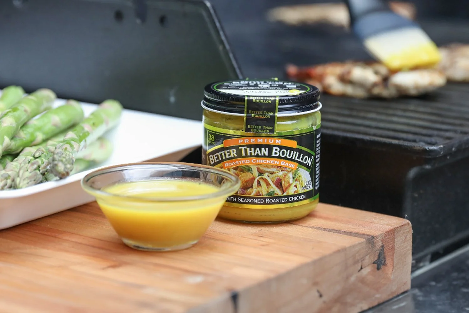 A jar of Better Than Bouillon Premium Roasted Chicken Base next to a small glass bowl of prepared broth on a wooden cutting board, with fresh asparagus and a grill in the background.
