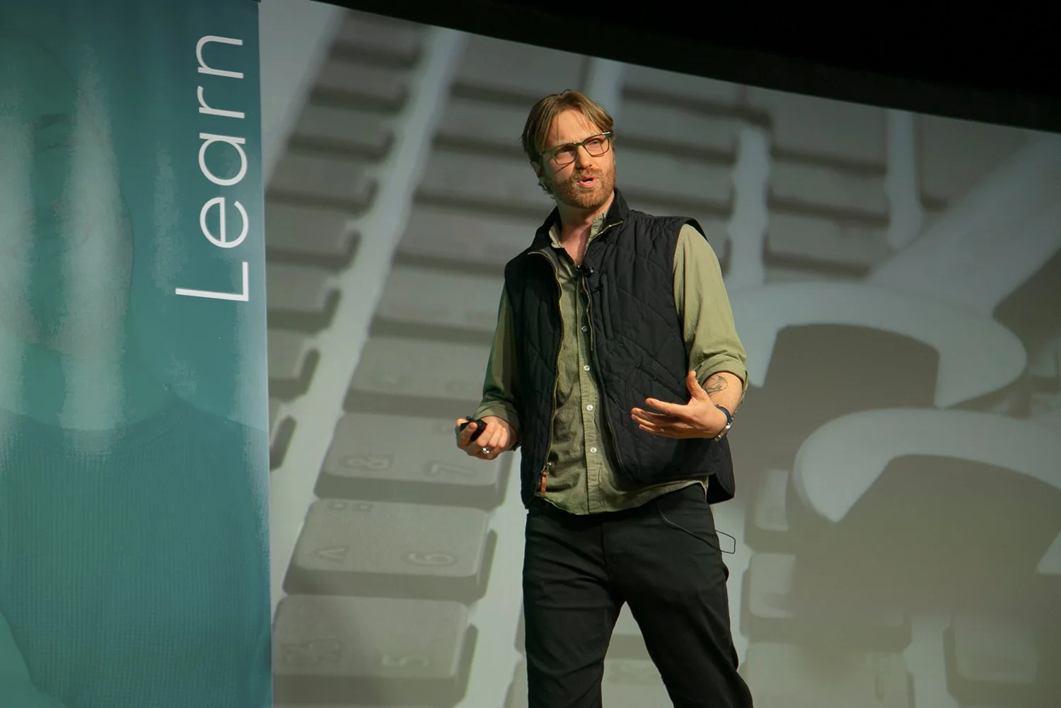 A man with glasses and a beard, wearing a green button-down shirt and a black quilted vest, gives a presentation on stage. Behind him is a large screen showing a close-up of a keyboard and the word 'Learn'