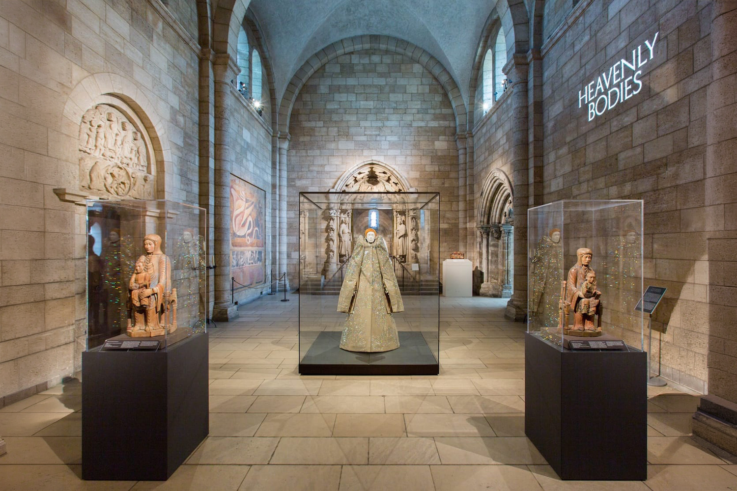 A gallery view of the 'Heavenly Bodies' exhibition at The Met Cloisters, featuring a large avant-garde ensemble centered between two medieval 'Throne of Wisdom' sculptures