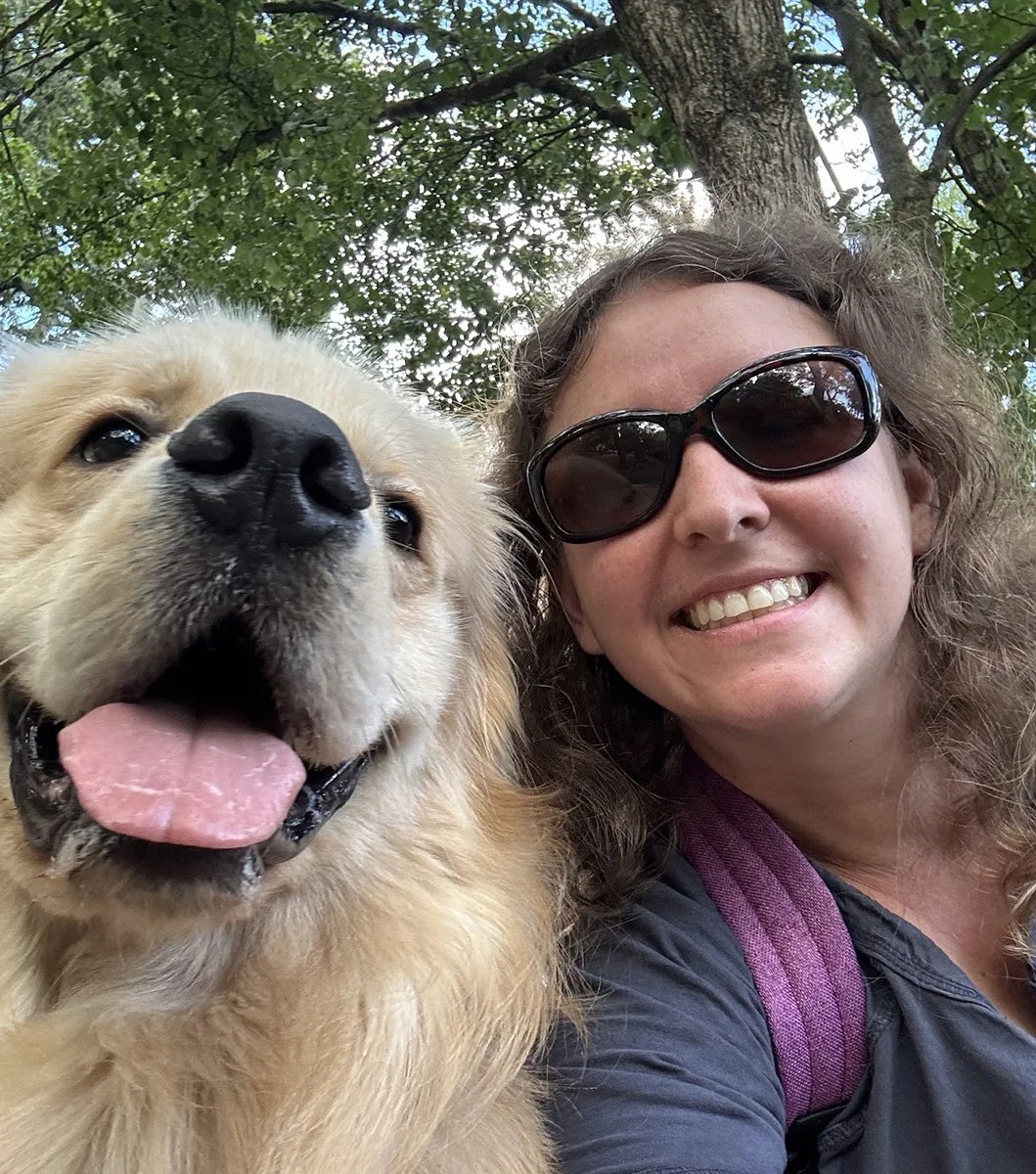 A woman with curly hair wearing sunglasses smiling next to a happy golden retriever dog outdoors under a tree.