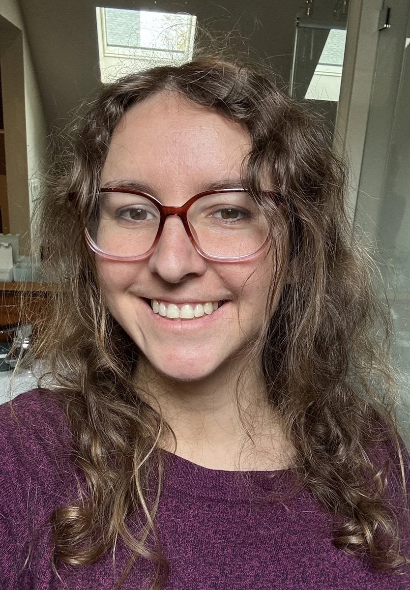 Photo of Annick, a woman with long hair wearing red glasses and a maroon sweater, smiling at the camera, indoors near a window.