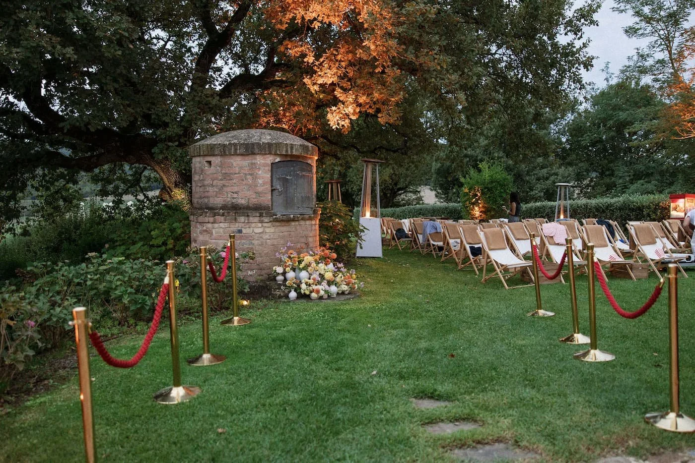 Outdoor open-air movie screening lounge on a lawn featuring wooden deck chairs with camel-colored blankets, red velvet rope stanchions, and a brick oven decorated with flowers at Rosewood Castiglion del Bosco