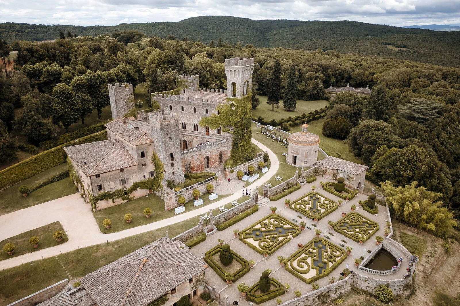 High aerial drone view of a luxury medieval castle wedding venue in Tuscany, featuring an ivy-covered stone tower, historic estate buildings, a small chapel, and a large, manicured Italian parterre knot garden surrounded by dense forests.