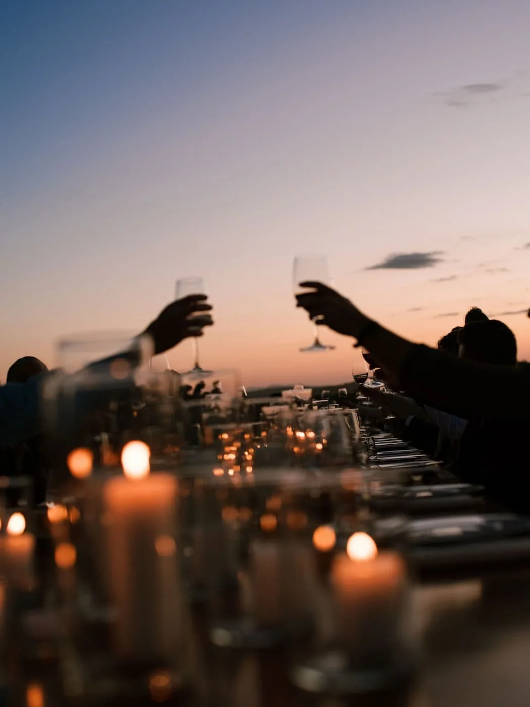 Silhouetted guests raising wine glasses in a toast above glowing candles along an outdoor dinner table at sunset at Rosewood Castiglion del Bosco.