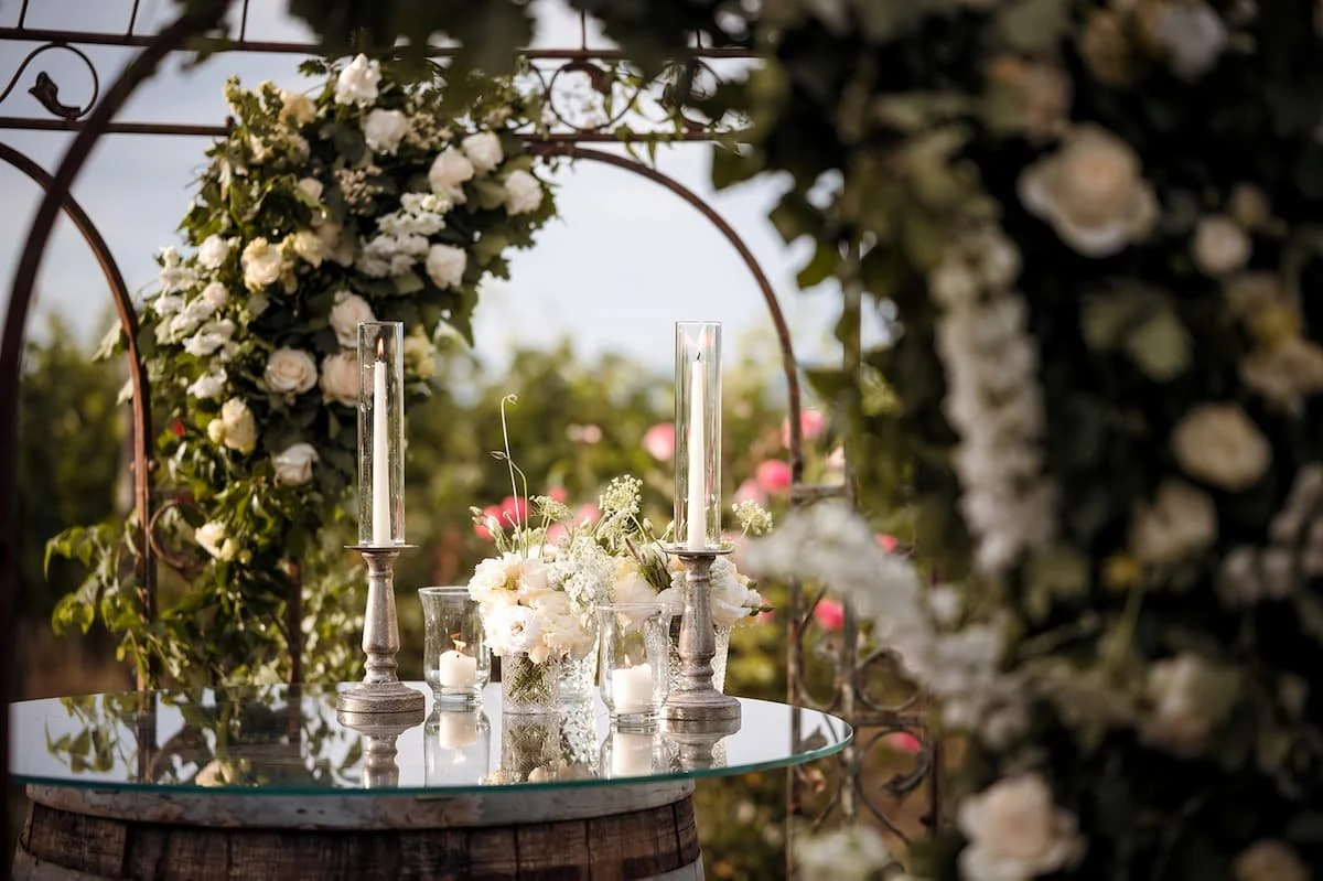 A romantic luxury proposal setup in a Tuscan vineyard featuring a rustic wine barrel table topped with a glass mirror, elegant white floral arrangements, and tall glowing candles framed by a white rose arch.
