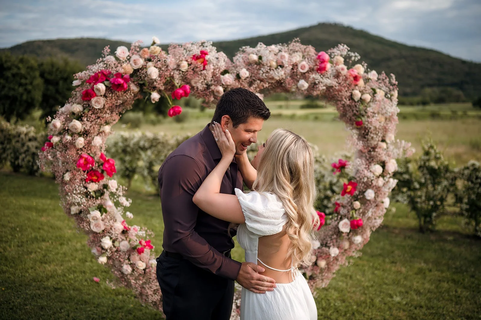 A smiling bride-to-be cradling a man's face as they celebrate an outdoor luxury elopement or wedding proposal in Tuscany, framed by a large heart-shaped floral arch made of baby's breath, pink roses and magenta peonies