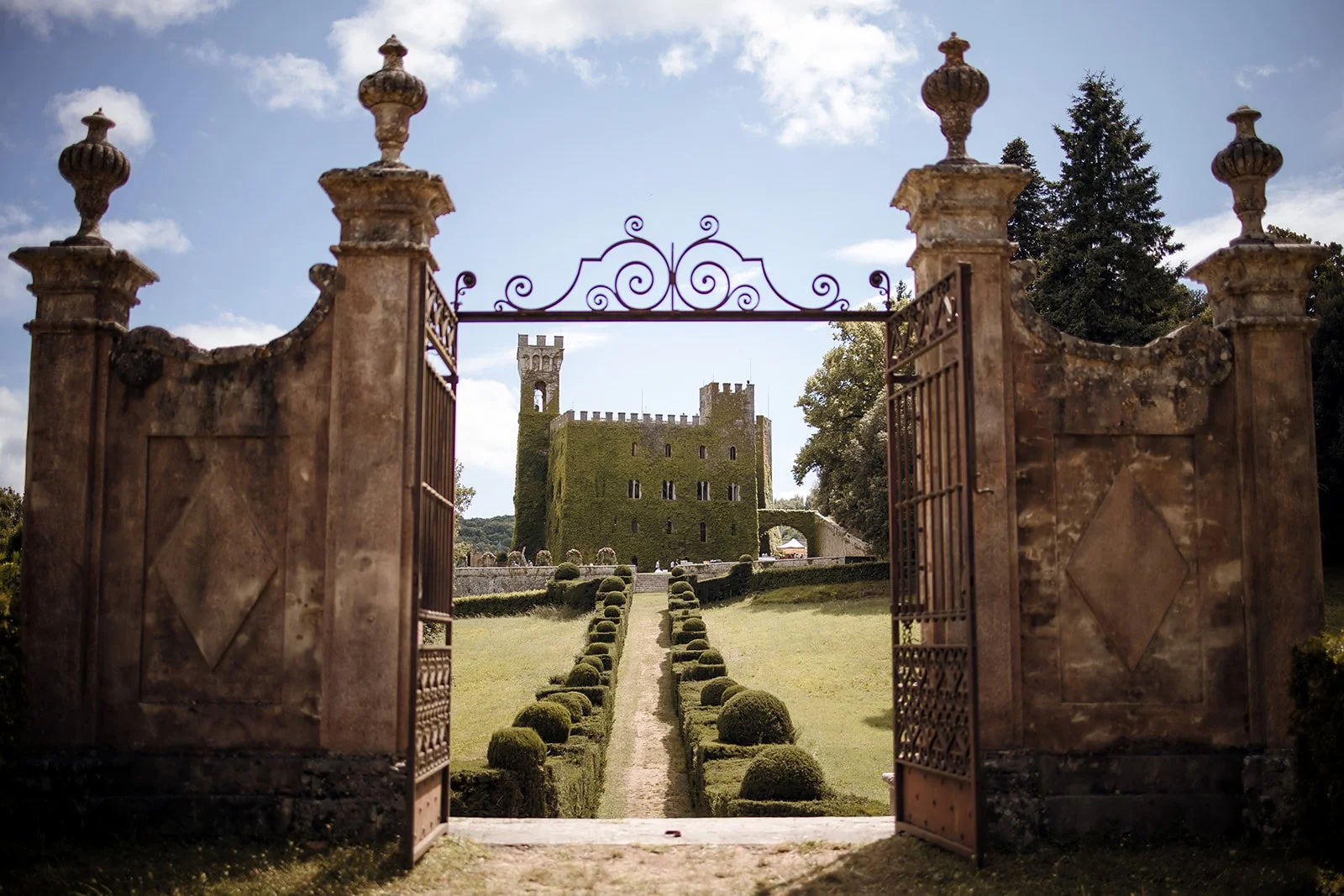 Looking through large historic stone entrance pillars and open wrought iron gates down a manicured boxwood hedge path toward a grand, ivy-covered medieval castle wedding venue in Tuscany under a blue sky.