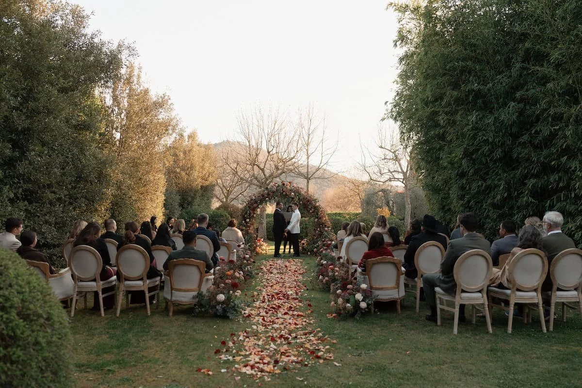 Outdoor winter wedding ceremony in Tuscany featuring a lush, circular floral arch, a petal-strewn aisle runner, and elegant round-backed chairs under crisp, clear skies with bare winter trees in the background.