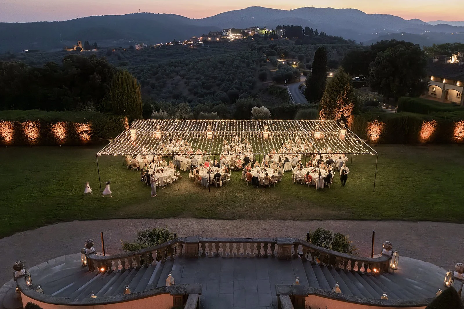 An elevated view looking down from a outdoor luxury wedding reception dinner in Tuscany, featuring round guest tables illuminated beneath a large fairy light canopy on a lush lawn with twilight views of distant olive groves and a hilltop village.