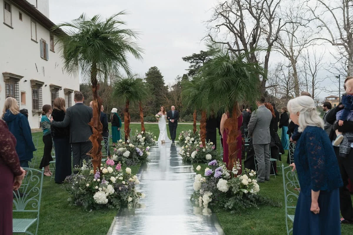 Bride walking down a reflective silver aisle flanked by palm trees and floral meadow arrangements at Villa Corsini.