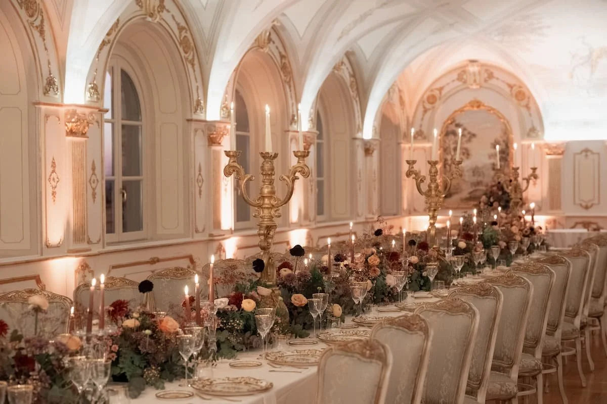 Grand indoor winter wedding reception banquet table in a luxury Tuscan villa, featuring ornate gold candelabras, tall lit taper candles, warm taupe and muted floral arrangements, and gilded antique dining chairs under vaulted ceilings.