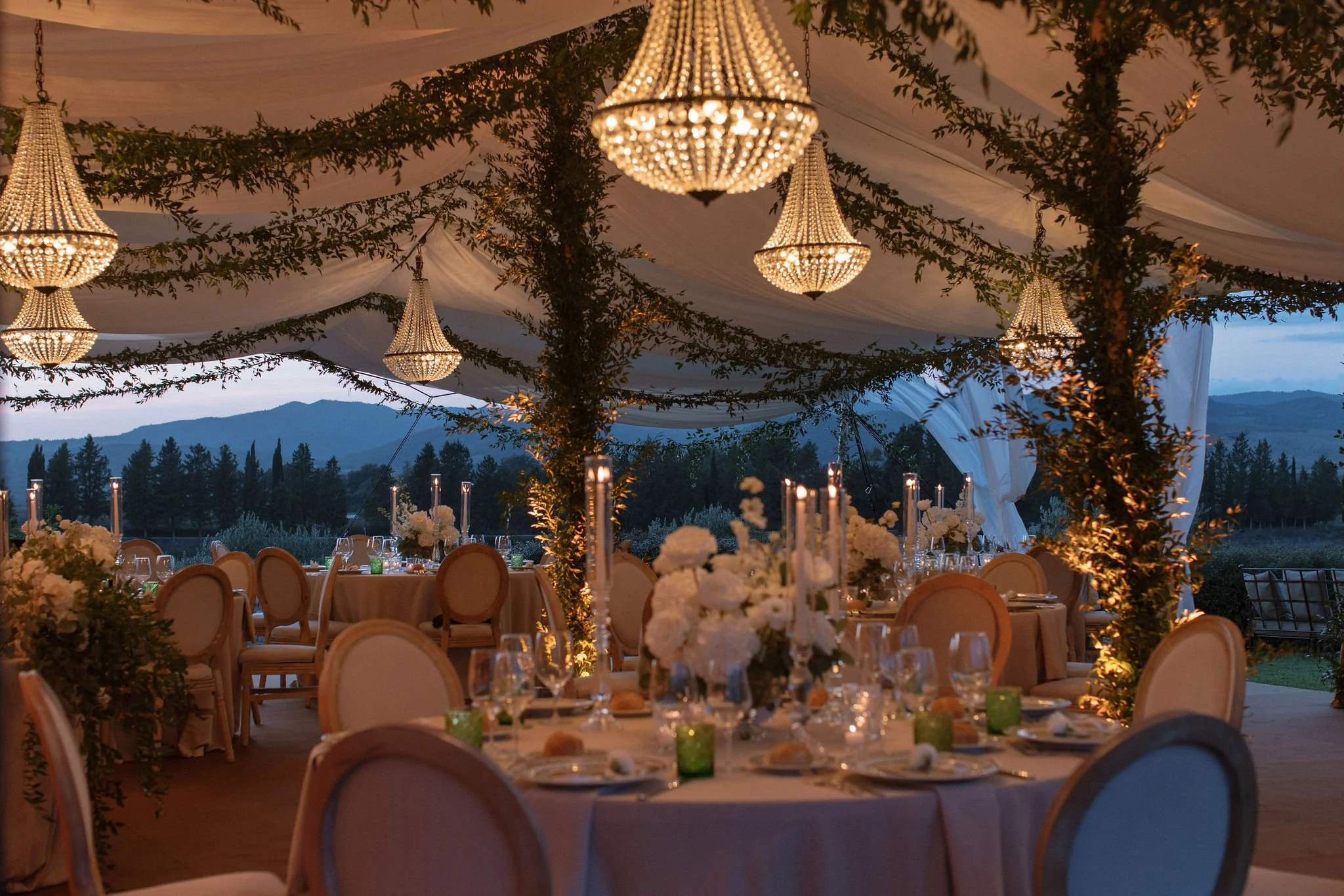 An elegant luxury wedding reception under a draped marquee marquee tent in Tuscany, featuring round tables with white floral centerpieces, crystal chandeliers, greenery-wrapped pillars, and twilight views of the Italian countryside.