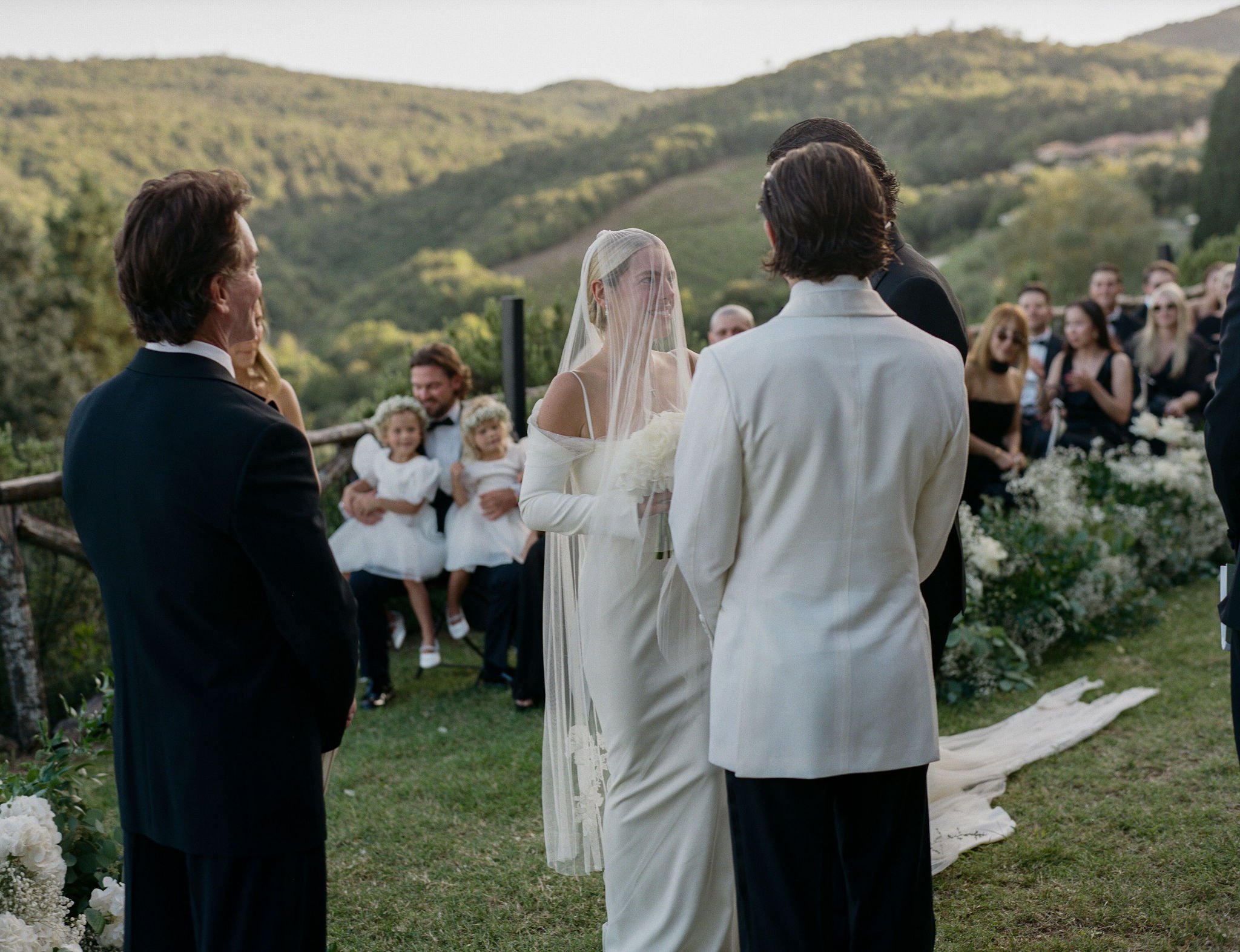 Bride in a sheer veil and elegant gown looking at her groom during an outdoor luxury wedding ceremony in the rolling hills of Tuscany, Italy.